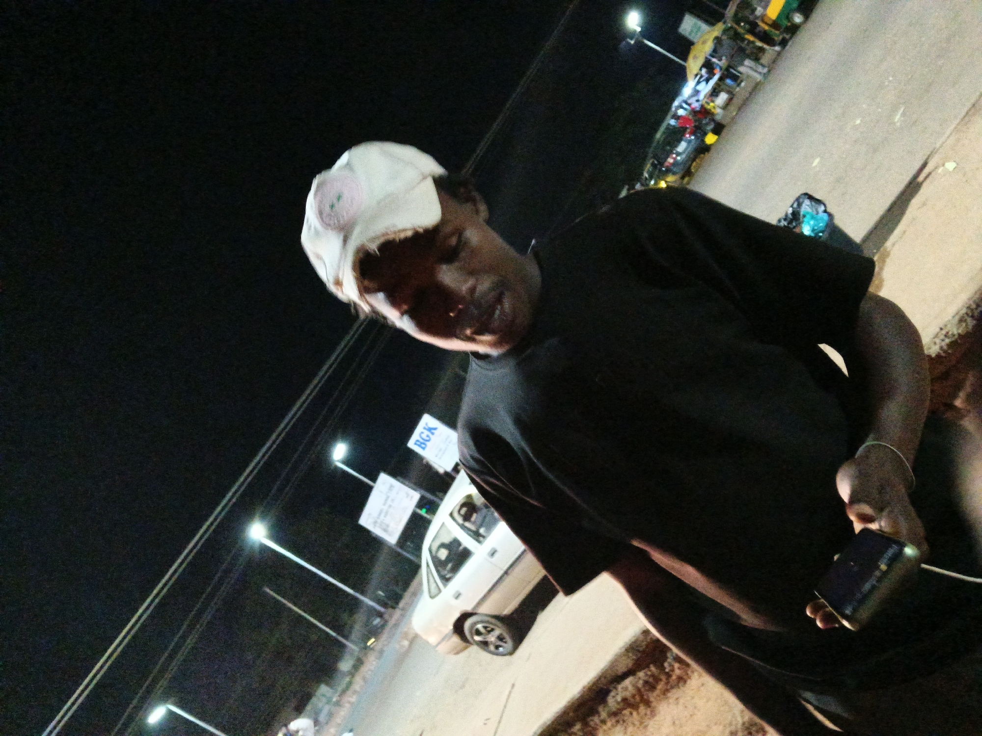 At night in Bukuru, Nigeria, a young man wearing a white baseball cap and a black t-shirt is standing outdoors. He is holding a dark-colored object, possibly a phone, in his right hand. In the background, several streetlights illuminate a road with traffic, including cars and what appears to be a bus. A white car is parked on the side of the road, and there are signs in the background, one of which displays the text "BGK." The ground is paved, and there is a shadow cast by the man on the ground, indicating the direction of the light source. The overall atmosphere is dark, typical of a nighttime urban environment.