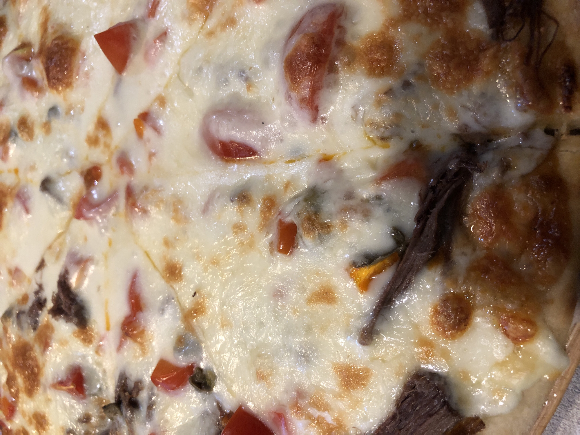 The image displays a close-up, top-down view of a baked pizza. The pizza features a generous layer of melted white cheese, exhibiting areas of golden-brown caramelization. Visible toppings include numerous irregularly diced red tomatoes, dark brown shredded or pulled meat, and small pieces of what appear to be green and orange/yellow bell peppers or similar vegetables interspersed beneath and within the cheese. The pizza has been cut into slices, with division lines evident across the surface. The crust edge, visible at the bottom and left, appears golden brown. No specific location within the city of Chaikovsky, Russia, nor any individuals, actions, or interactions, are discernible from the image content.