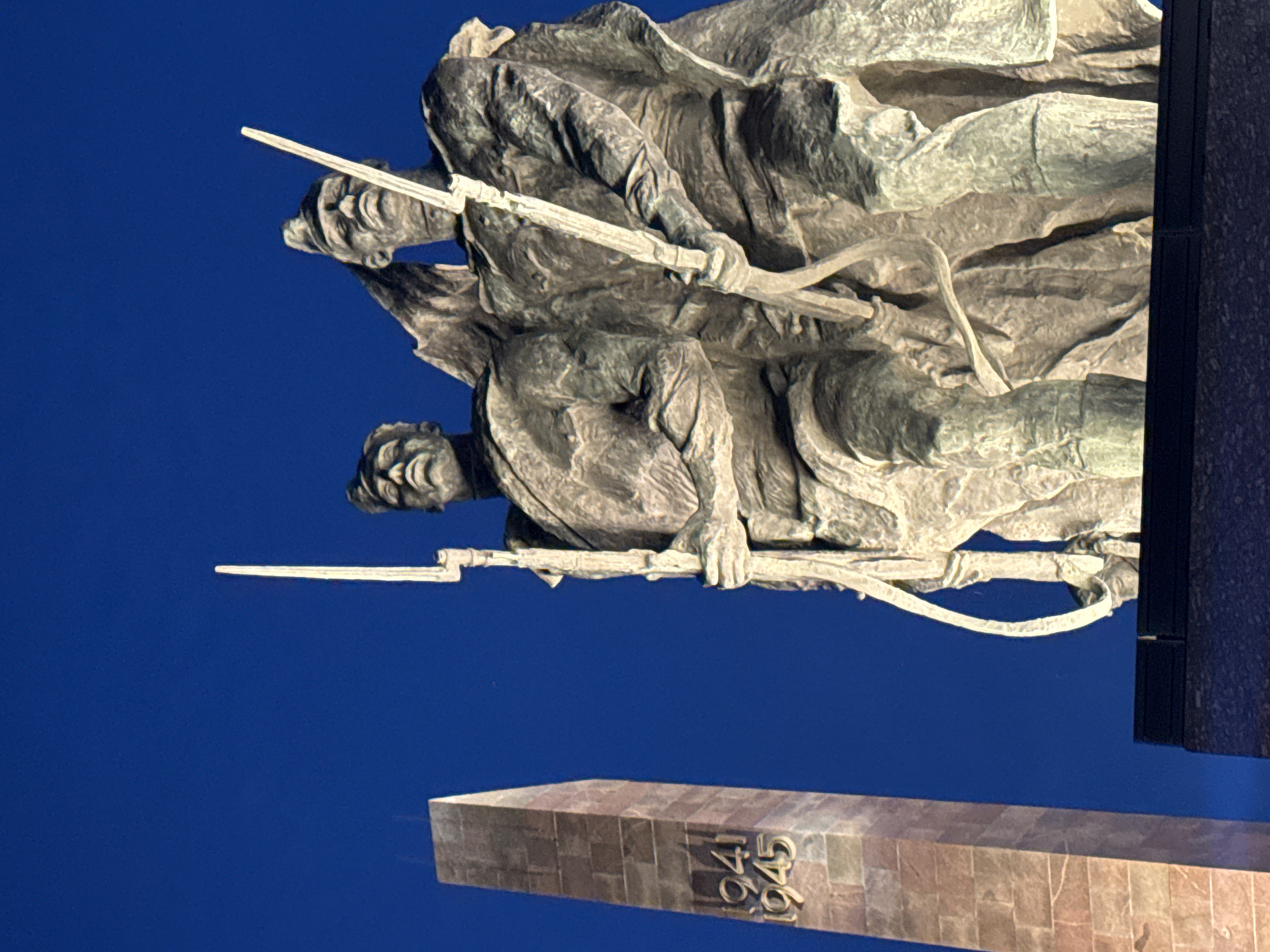 The image displays a low-angle view of a monumental war memorial under a deep blue sky, likely at twilight or night with artificial illumination. In the foreground and center, two heroic-scale bronze statues of male soldiers stand prominently. Both figures are depicted holding long, vertical rifles or bayonets. The soldier on the left is positioned slightly forward of the soldier on the right, and both gaze upwards with stern expressions. Their attire appears to be military uniforms. To the left, a tall, light-colored, rectangular stone obelisk is visible, with the years "1941" and "1945" inscribed vertically on its visible side. The base supporting the statues is dark and textured, occupying the bottom edge of the frame. This scene depicts a section of the Monument to the Heroic Defenders of Leningrad, located at Victory Square (Ploshchad Pobedy) in Saint Petersburg, Russia.