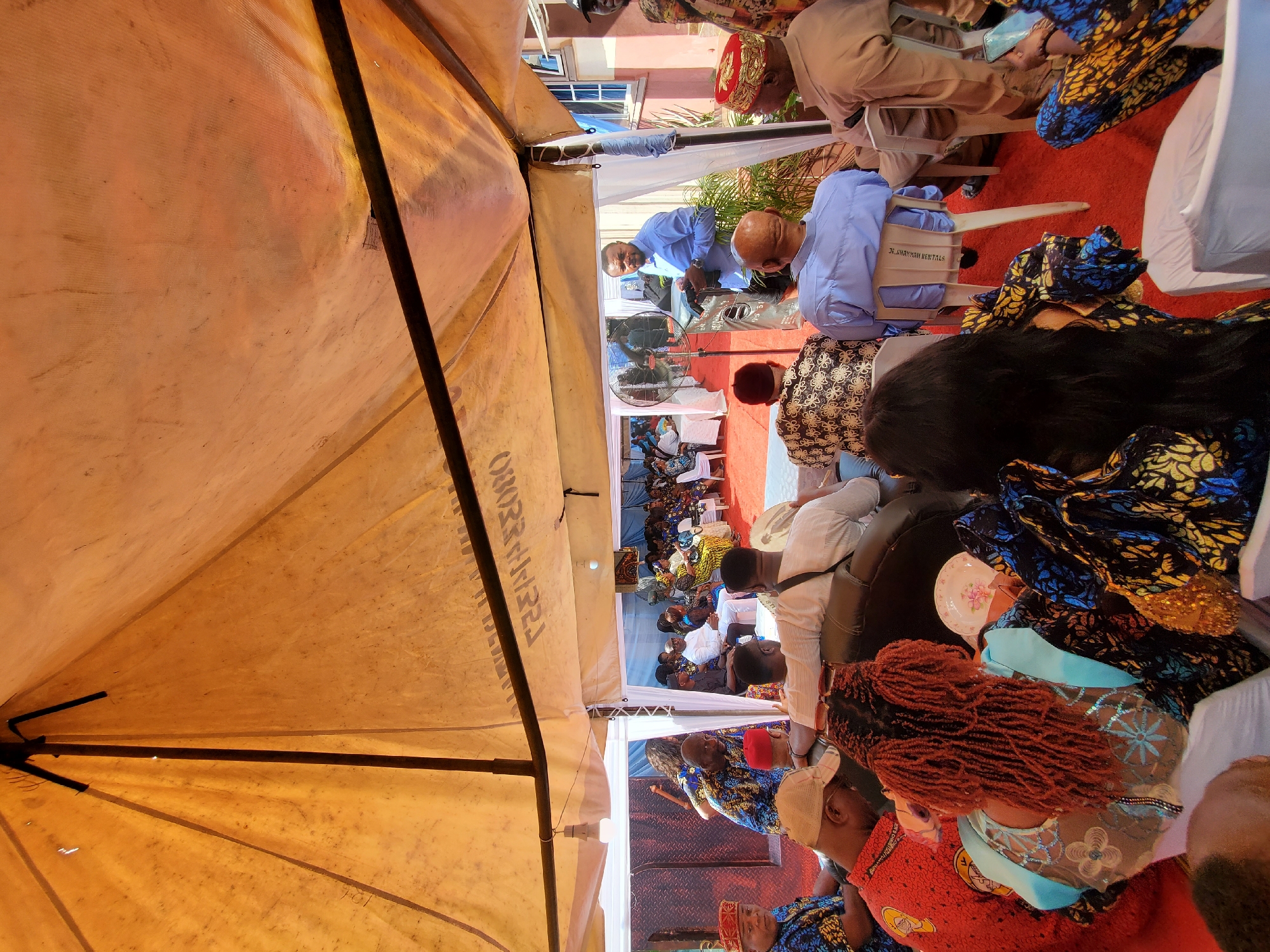 The heavily rotated image depicts an outdoor social gathering or event in Obosi, Nigeria, viewed from within a large, somewhat aged, orange-brown tent. The tent's interior fabric and dark support poles fill the immediate foreground and left side of the frame. Underneath this tent and extending to adjacent white canopy tents, numerous people are seated on plastic chairs arranged on a red carpet.
In the midground, several individuals are visible. A man in a light blue collared shirt is seen from the front, seemingly addressing or interacting with someone, while another man in a similar blue shirt is viewed from behind, seated on a beige plastic chair. The back of this chair clearly displays the text "J.R. SHANNAH RENTALS". A large standing table fan is positioned nearby, suggesting an effort to provide comfort from the heat. Many attendees are dressed in colorful traditional African patterned attire. In the foreground right, a woman with prominent reddish-brown dreadlocks is seen from behind, wearing a light blue embroidered top and a red patterned fabric. A small white plate with a floral design rests near her.
Further in the background, more guests are seated under additional white tents, with a light-colored building visible beyond. The scene conveys a sense of a formal or celebratory gathering, taking place during the daytime. Faint, partially obscured text, possibly "RENTALS," is also visible on the interior fabric of the large orange tent.
