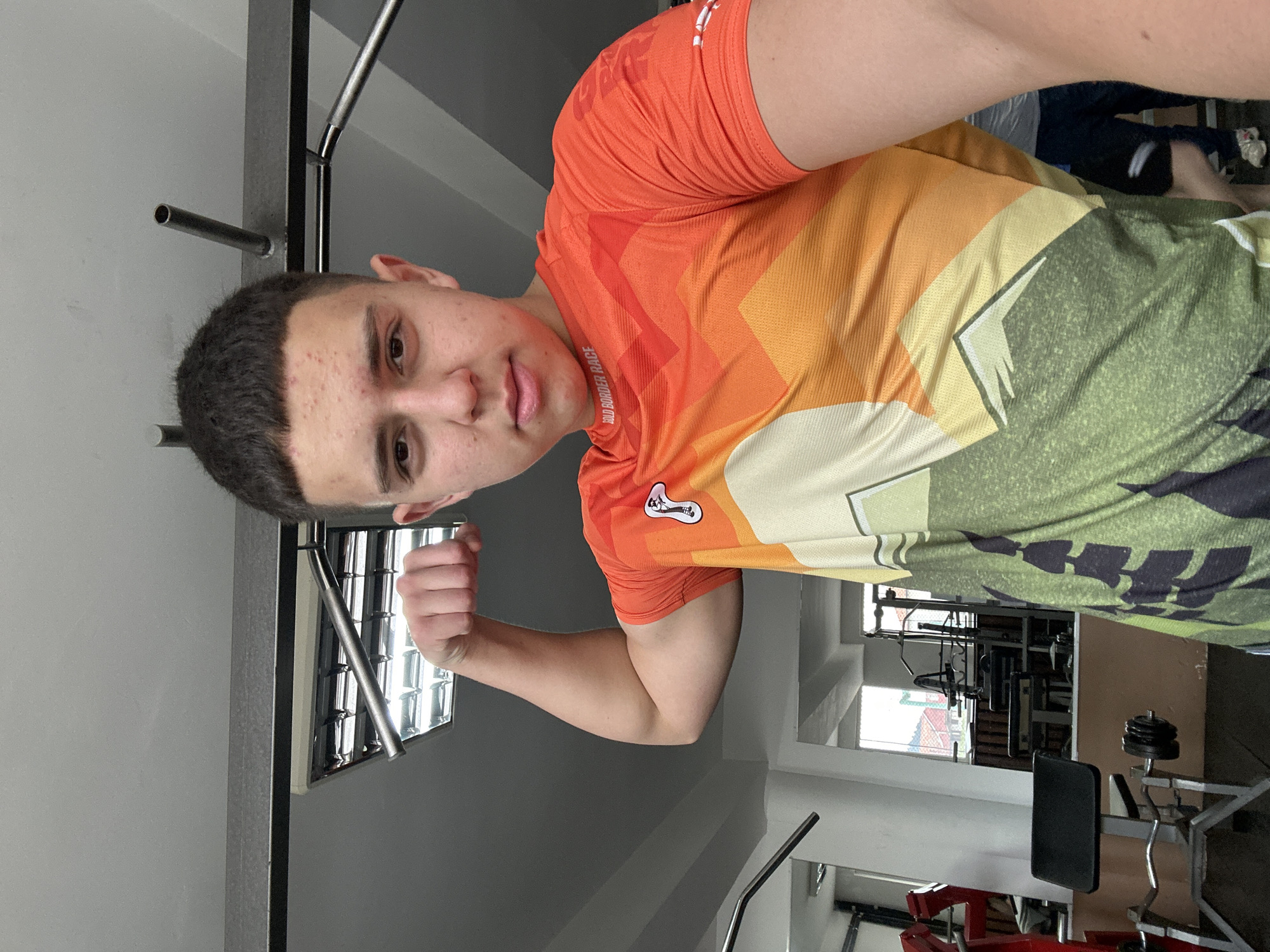 A young man is photographed in a gym. He is wearing an orange, yellow, and green shirt and is flexing his left arm. The gym has workout equipment, including weights and exercise machines. The lighting is provided by fluorescent ceiling lights. The location is Zlatograd, Bulgaria.