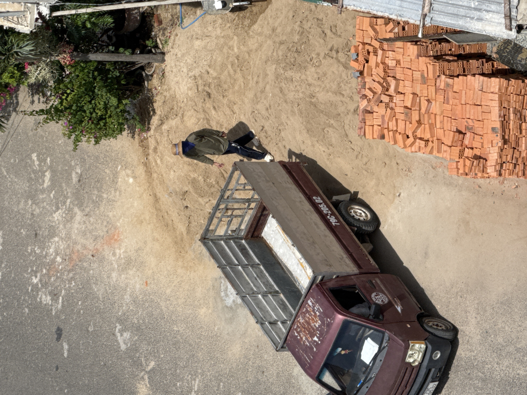 In Bac Nha Trang Ward, Vietnam, an overhead view captures a construction scene. A maroon flatbed truck with a metal cage is parked on a dusty surface next to a pile of orange bricks. A person wearing a straw hat, a green long-sleeved shirt, and dark pants walks away from the truck, heading towards the pile of bricks. The ground is a mixture of asphalt and sand, with green foliage and a palm tree visible in the background, alongside a building.