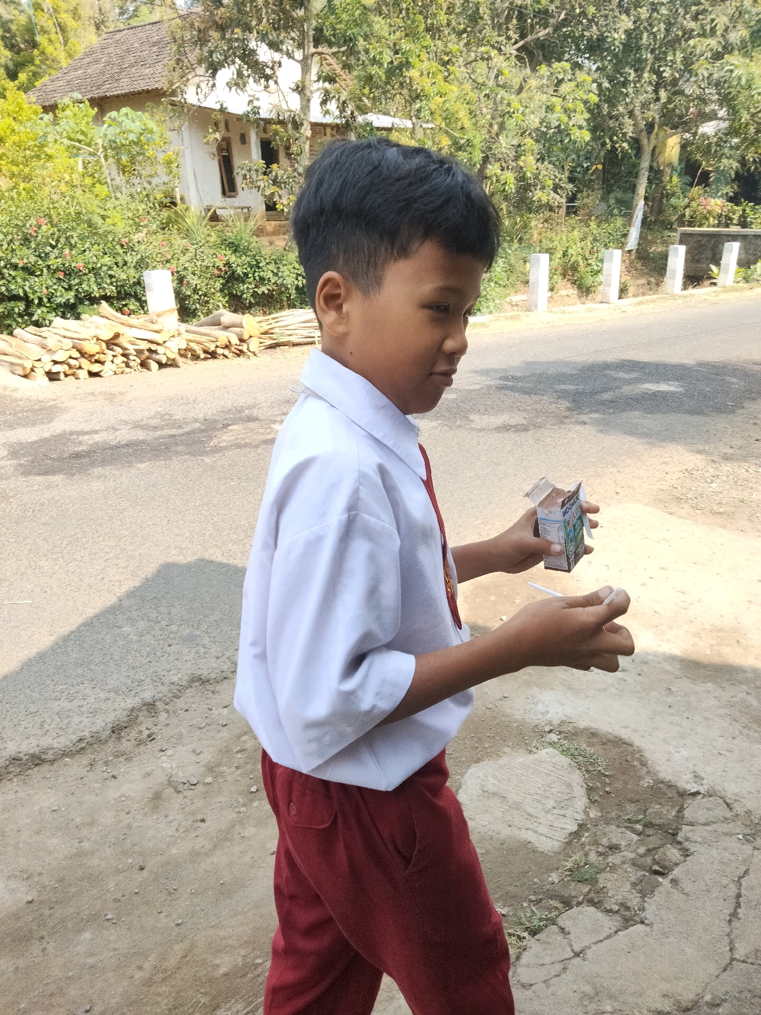 A young boy in a white school uniform shirt and red pants stands on a dirt and asphalt surface, holding a small chocolate milk carton. He is looking to his right with his mouth slightly open. In the background, a white house with a dark roof is partially visible, surrounded by lush green vegetation and trees. A pile of cut wood is stacked on the left side of the road. Several white concrete posts are lined up along the side of the road. The lighting suggests it is daytime. The setting appears to be a residential area in Sidorejo, Indonesia.