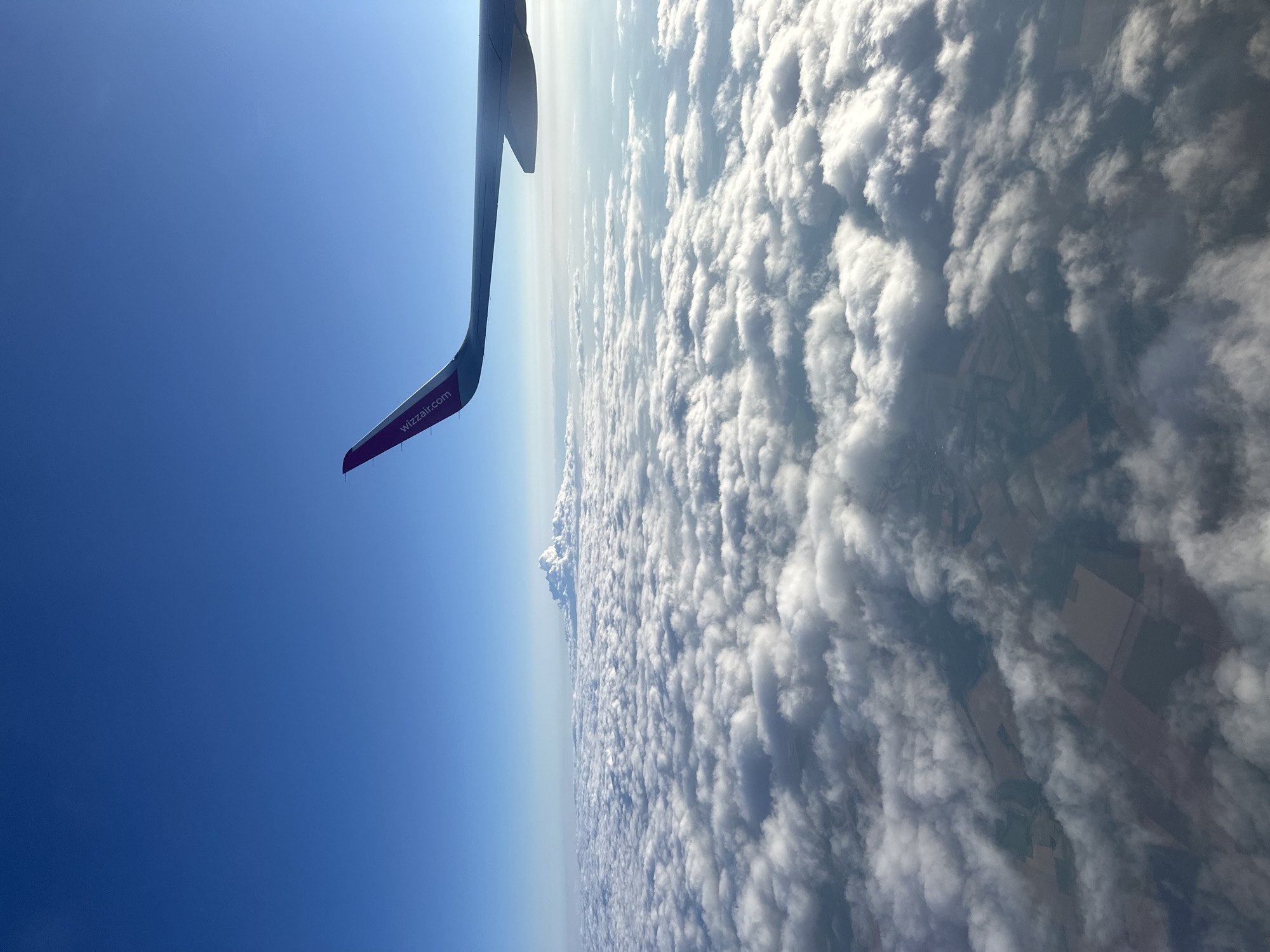 An aerial view from an airplane window shows a vast expanse of white clouds stretching across the horizon. The wing of the aircraft, with "wizzair.com" printed on it, extends into the upper right corner of the frame. Below the clouds, hints of agricultural fields and land are visible, suggesting a rural or semi-rural landscape. In the distance, a towering mountain range, likely the Balkan Mountains, is partially obscured by a large, cauliflower-like cloud formation. The sky is a clear, deep blue. The image captures the scene above the city of Vekilski, Bulgaria.