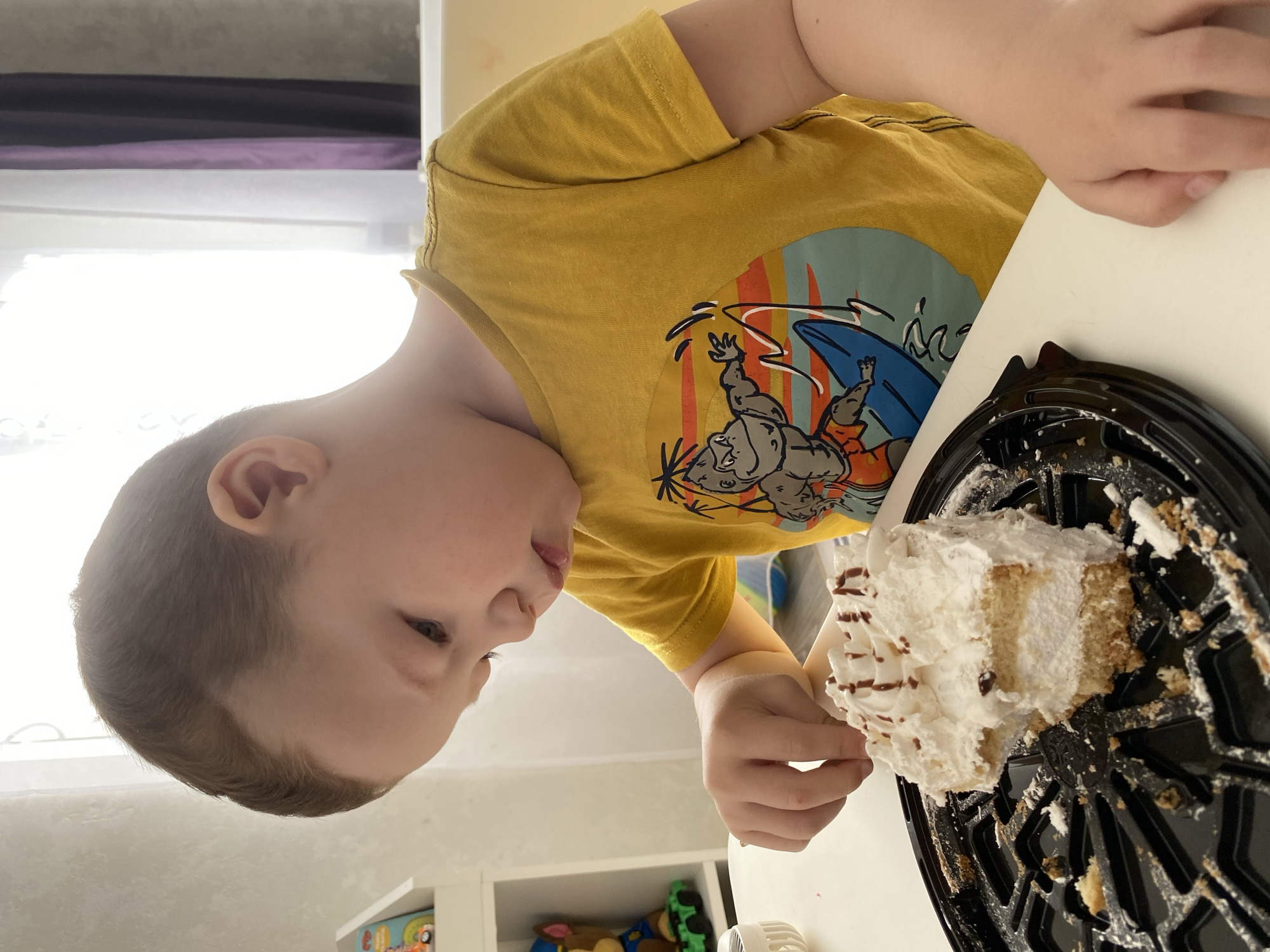 A young boy in a yellow shirt is eating a piece of cake with white frosting. The cake sits in a black plastic container. The location is indiscernible and cannot be confirmed as Sochi, Russia.