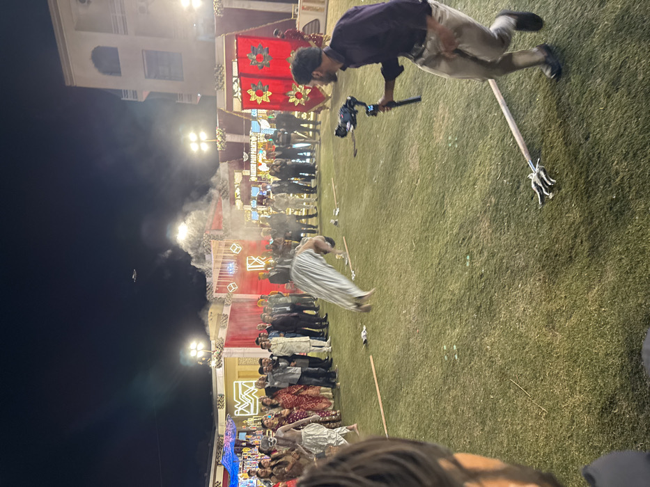 An outdoor celebration unfolds at night, with an elaborately decorated stage serving as the backdrop for a gathering of people. Smoke or fog drifts in the foreground, adding to the festive ambiance. A man wearing a gray shirt and khaki pants bends over, holding a camera stabilizer. Scattered on the grass are wooden staffs, some with metal trident-like attachments. The scene suggests a cultural event or performance, captured from a slightly tilted perspective.