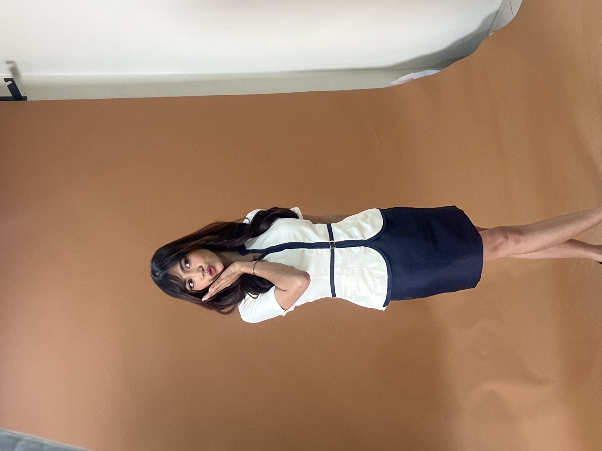 In a studio setting with a plain brown backdrop, a young woman with long, dark hair is posing. She is wearing a white short-sleeved top with a dark blue trim and a matching dark blue skirt that ends above her knees. Her left hand is placed on her cheek, and her lips are pursed in a playful gesture. A thin, dark belt with a buckle is cinched at her waist. To the right of the woman, a white backdrop is partially visible, suggesting a professional photography setup. There is no visible indication of the specific location within Davao City, Philippines.