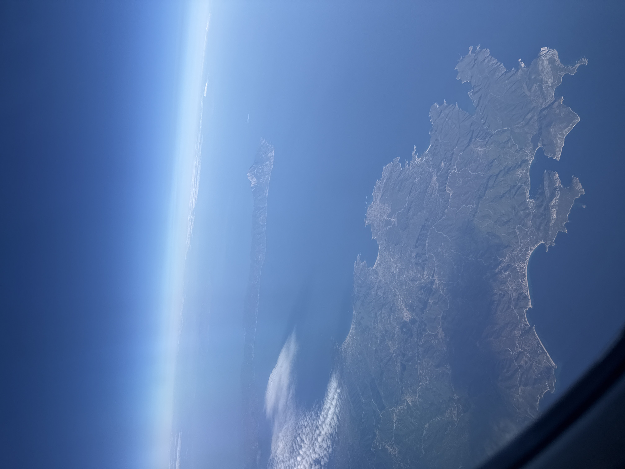 This is an aerial photograph taken from an airplane, looking down at a coastline. The water is a deep blue, and the land is a mix of green and brown, with visible signs of human habitation such as roads and buildings. There are some clouds in the sky, and the horizon is visible where the atmosphere meets space. The image does not contain any people, objects, or actions that would allow for identification of the city of Lukovit, Bulgaria, or any specific location.