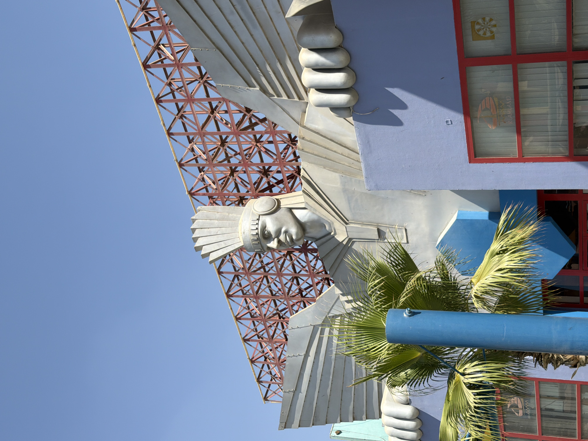 The image shows a close-up, low-angle shot of a large, metallic statue of a stylized human figure with feathered wings and headdress, possibly representing an ancient deity or historical figure. The statue is located in the city of Sheikh Zayed, Egypt, as indicated by the text "REALAND" visible through a window on the building facade. The statue is made of a silver-colored material and has a strong, imposing presence. Its features are sharp and angular, with a distinct headdress, ear adornments, and large hands. Behind the statue, a red metal lattice structure is visible against a clear blue sky. The building the statue is attached to is painted a light purple and features red-framed windows with white blinds. A palm tree with vibrant green fronds partially obscures the lower part of the statue and the building. The overall scene suggests an outdoor architectural feature or a monument in a public space within the city.