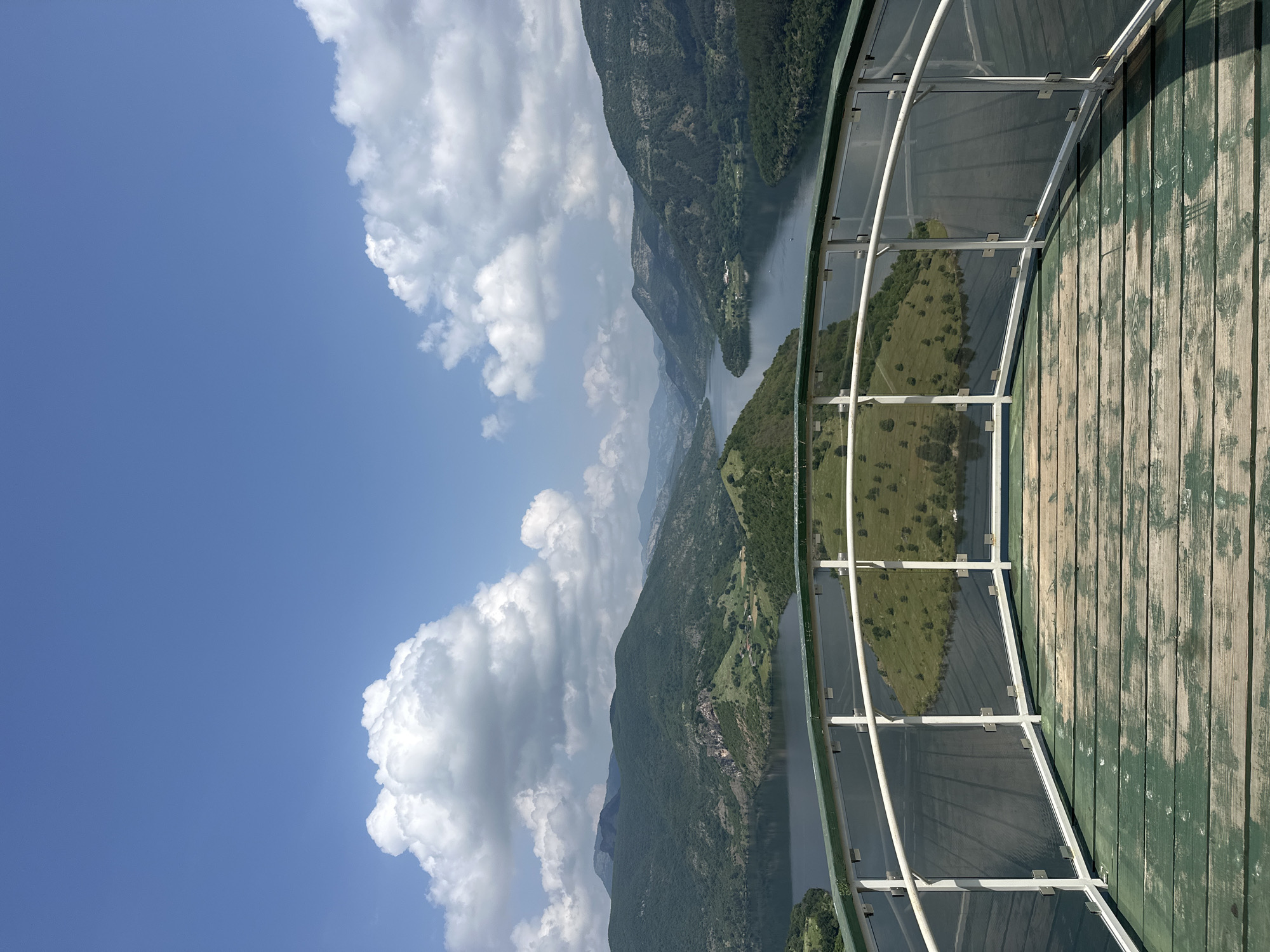 This panoramic image captures a breathtaking mountainous and waterscape view from an elevated observation deck in Borovitsa, Bulgaria.
In the foreground, a curving viewing platform is visible, featuring a floor constructed from weathered wooden planks, distinctly painted in a muted green hue. A modern safety railing, composed of white metal frames supporting clear glass panels, encircles the platform's edge, offering an unimpeded vista of the landscape.
Beyond the railing, a serene, expansive body of water, likely a reservoir or a wide river, stretches across the midground. Its surface appears calm, subtly reflecting the bright sky. Within this water, a prominent, irregularly shaped island or peninsula stands out, covered in lush green grass and dotted with numerous small trees.
The background is dominated by a series of majestic, heavily forested mountains that recede into the distance. Their verdant slopes and peaks exhibit varying shades of green and blue-grey, indicating depth and scale. Overhead, the sky is a vivid blue, populated by large, fluffy white cumulus clouds that add texture and drama to the scene.
The image portrays a clear, bright day, likely late morning or early afternoon, with sunny weather. No people or visible text are present, emphasizing the untouched natural beauty and grandeur of the mountainous landscape. The elevated perspective conveys a sense of peaceful solitude and vastness.