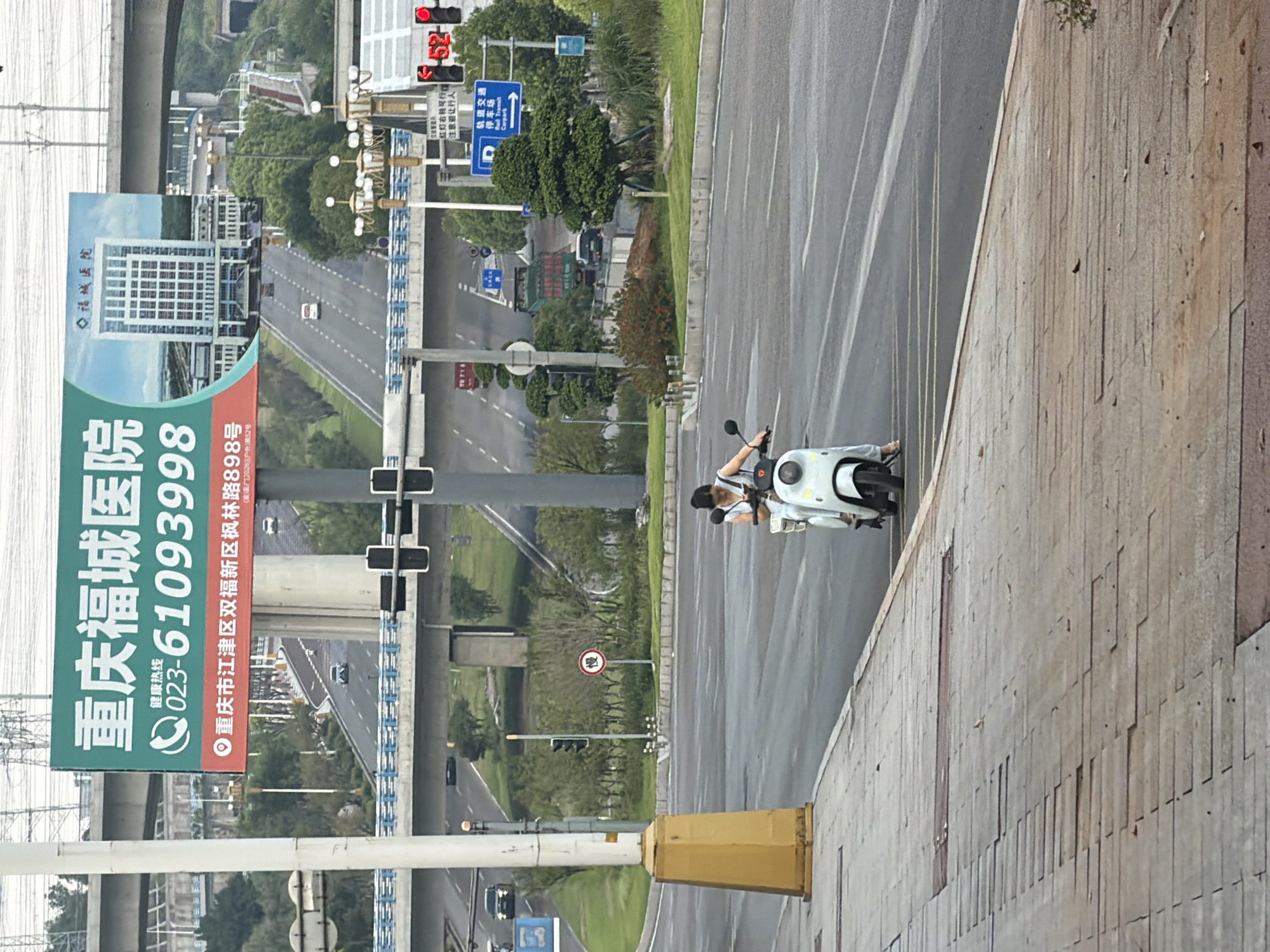 In a city street scene, a woman rides a white electric scooter on the right side of the road, moving away from the viewer. She is wearing a light-colored top and light pants. The scooter has a visible front light and is moving on a paved road with yellow lane markings. The sidewalk in the foreground is made of gray and red paving stones, with some sections appearing uneven or broken.
In the background, a large green billboard displays information about "Chongqing Fucheng Hospital," including a phone number "023-61093998" and an address in Jiangjin District. The billboard also features an image of a modern hospital building. Beyond the billboard, elevated highways and bridges are visible, with cars and trucks moving on them. Traffic lights and street lamps are also present. The overall environment suggests a busy urban area in China, likely within the city of Chongqing, specifically the Jiangjin District as indicated on the billboard.