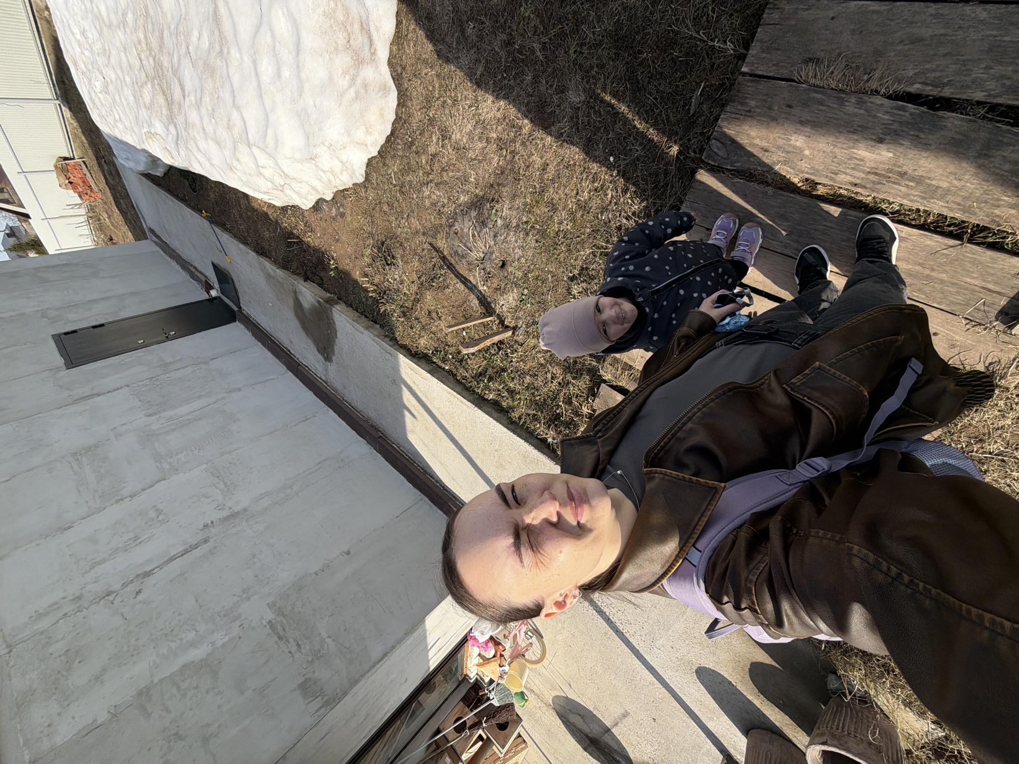 A man takes a selfie outdoors with a young child in Chaikovsky, Russia. A large pile of melting snow is visible on the grass next to a building with a flat roof. The man is wearing a brown leather jacket and a blue backpack. The child wears a dark jacket with a light hat and purple shoes. The ground is a mix of grass and wooden planks.