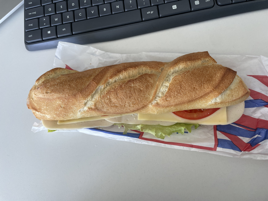 An overhead image depicts a baguette sandwich positioned on a white, unadorned surface, likely a desk, within Montigny-le-Bretonneux, France. The sandwich lies horizontally, slightly angled towards the top right. It consists of a golden-brown, crusty baguette split lengthwise, revealing visible fillings: green ruffled lettuce, several slices of light-colored deli meat (such as turkey or ham), a slice of light yellow cheese, and a red tomato slice. The sandwich is partially encased in white paper packaging, which displays a red and blue striped pattern on its left side. To the immediate right of the sandwich, a portion of a dark-colored computer keyboard is visible, featuring keys such as 'm', 'l', 'ctrl', 'alt', 'f11', and 'f12', consistent with an office or personal computing environment and likely an AZERTY layout. No individuals or active interactions are visible.