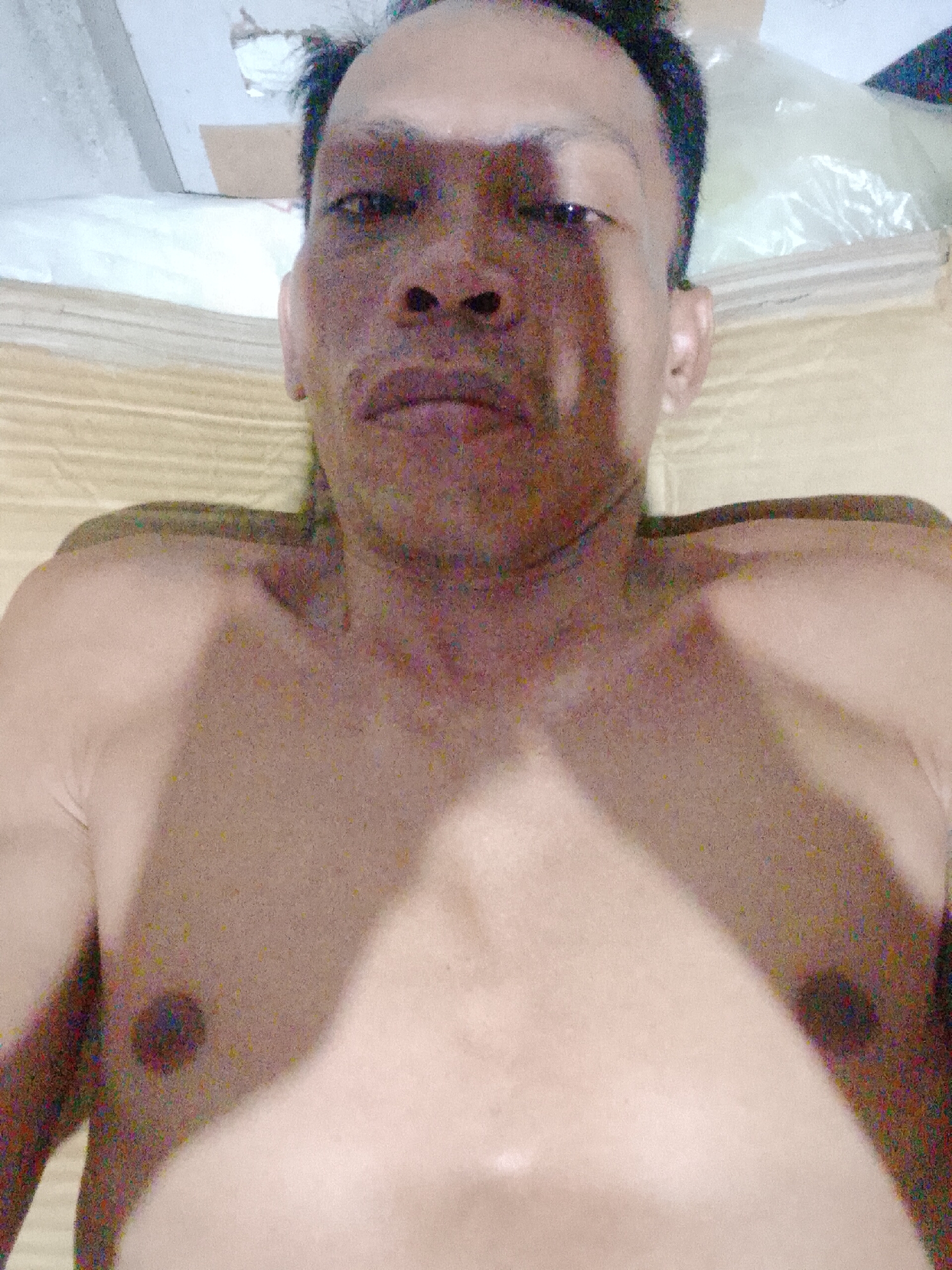 A shirtless person lies on a textured surface, possibly a mattress or blanket, with their head oriented towards the top of the frame. The person has dark hair and appears to be looking directly at the camera. The lighting in the scene is uneven, casting shadows across the person's face and torso. The background is blurry and indistinct, but possibly includes a white sheet or plastic bag. The specific location in Bukit Ketok, Indonesia, is not discernible from the image alone.