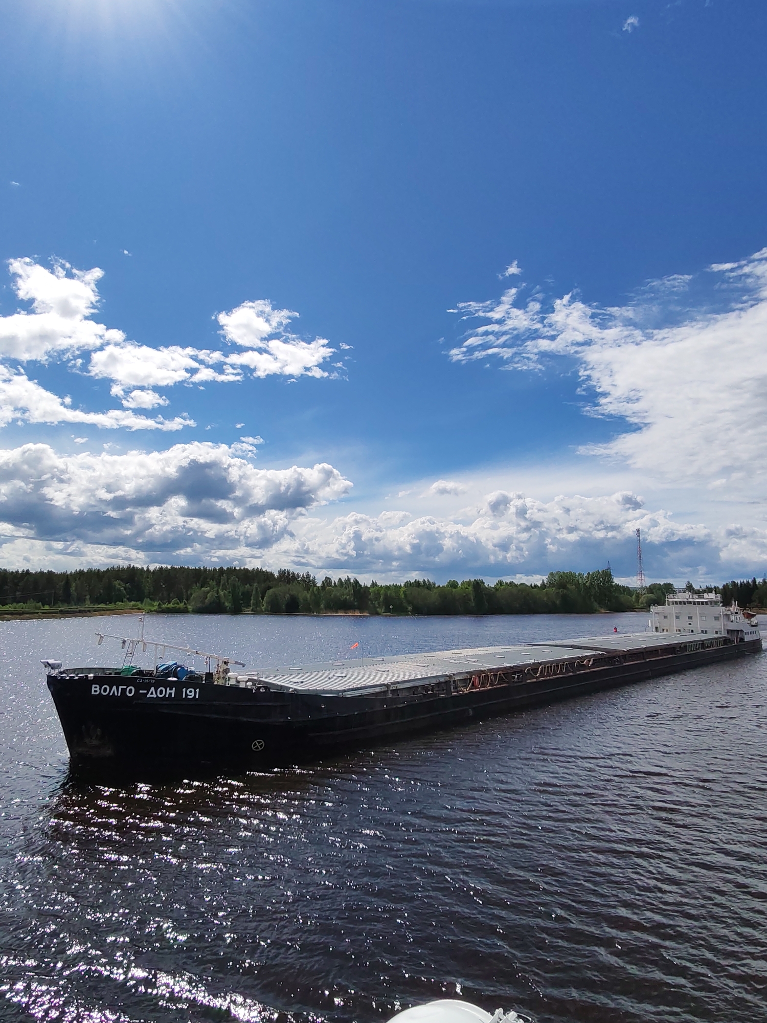 A large, dark barge labeled "ВОЛГО-ДОН 191" is on a body of water with reflections on the surface. Trees line the distant shore. The sky is bright blue with cumulus clouds. The image appears to be taken in daylight. Based on the label and the style of the ship, the location may be the city of Белоусово, Russia.