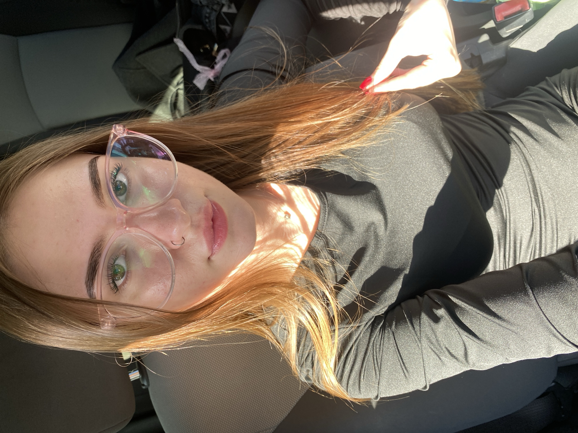 A young woman with long, auburn hair is pictured in a car, likely in Russia given the location context. She is wearing a black long-sleeved shirt and clear, pink-rimmed glasses. Her green eyes are visible, and she has a nose ring. Sunlight streams into the car, creating strong shadows and highlighting her hair and skin. One of her hands, with red-painted fingernails, is near her face, touching her hair. The interior of the car is visible, with a car seat and some bags in the background. The image appears to be taken during the daytime, with bright sunlight suggesting fair weather. The overall mood is casual and relaxed. There is no visible text in the image.