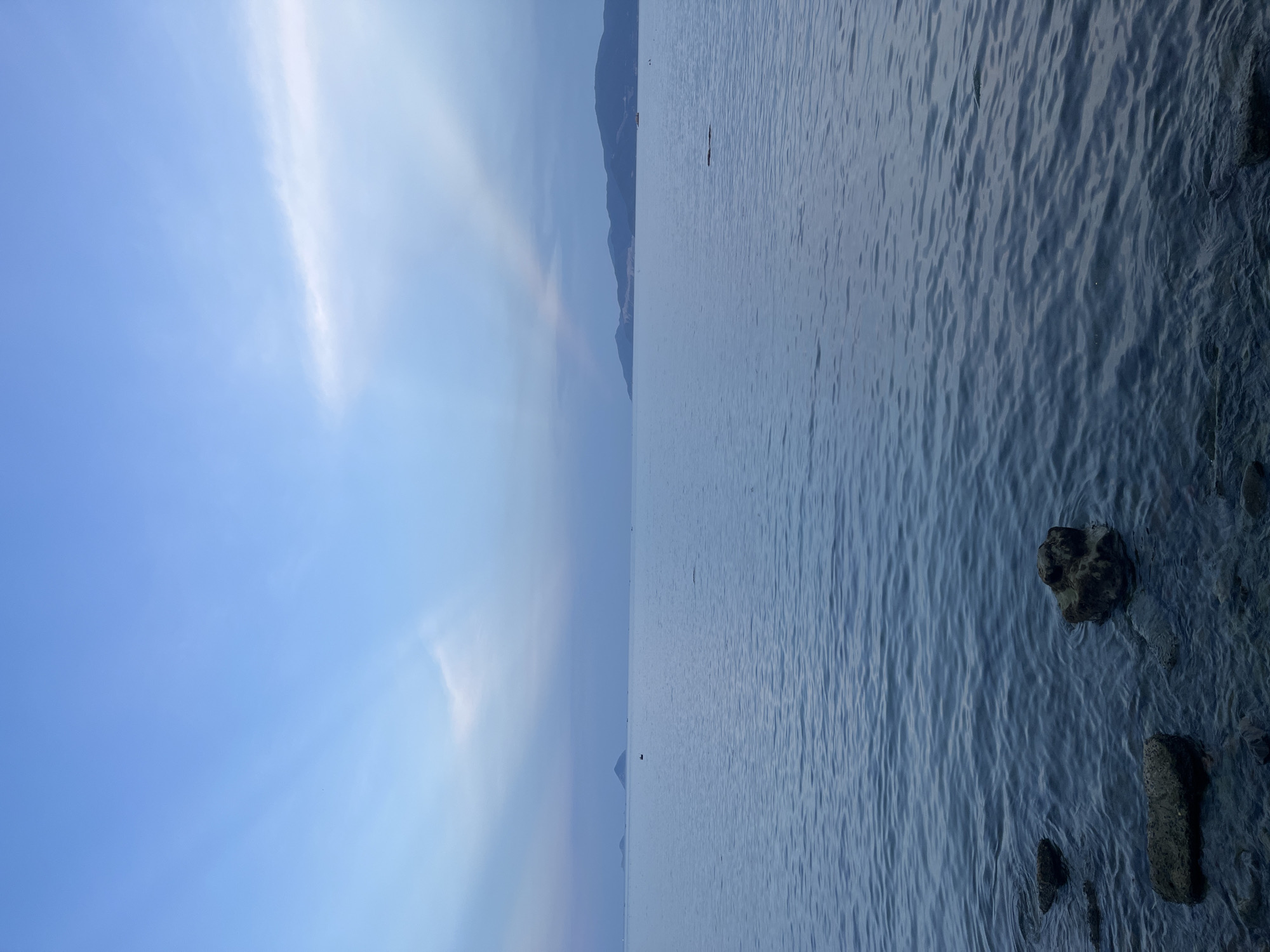 The image depicts a waterscape, likely a seascape, under a blue sky with wisps of clouds. A distant mountain range is visible on the horizon. The water appears calm with subtle ripples. In the foreground, rocks are partially submerged at the water's edge. Given the prompt's indication, this scene is likely in or near Bac Nha Trang Ward, Vietnam. No people are visible in the image.