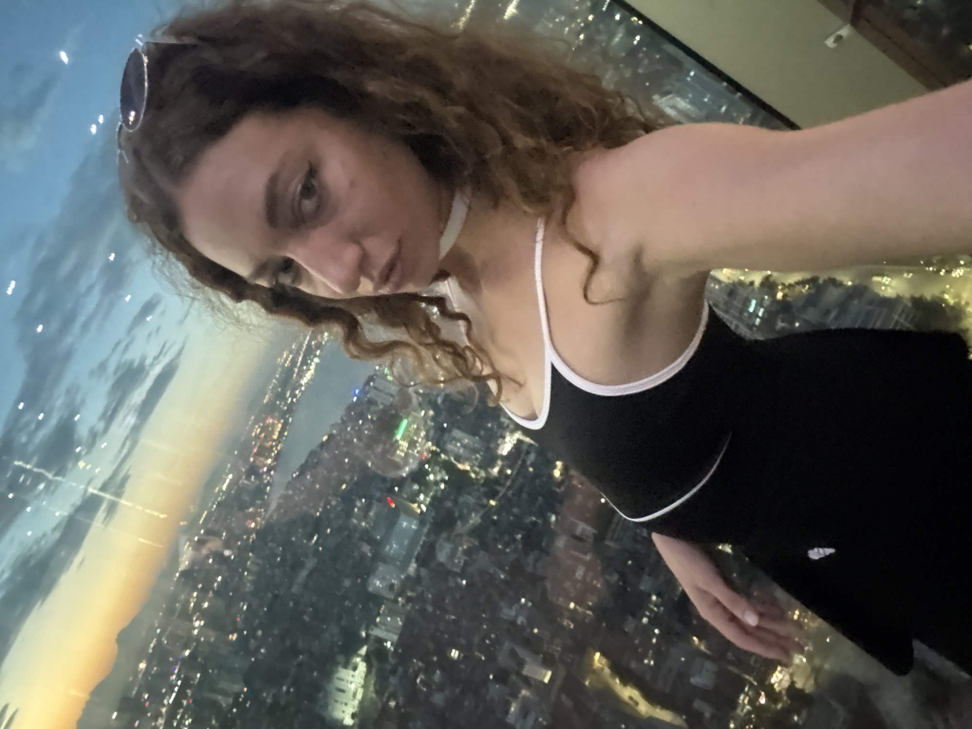 A woman is taking a selfie from a high vantage point, overlooking a city at dusk. She has curly brown hair and is wearing sunglasses on her head and a black top with white trim. A white choker necklace is visible around her neck. The city below is illuminated with lights, suggesting it is nighttime or twilight. The sky is a blend of blue and orange hues, indicating sunset or sunrise. The woman's expression is neutral. The scene appears to be from a tall building or observation deck, offering a panoramic view of the urban landscape. There is no visible text in the image.