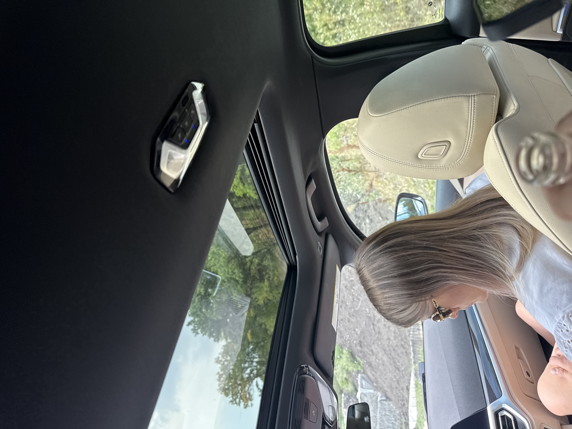 The image shows the interior of a car with a sunroof open. A woman with blonde hair and sunglasses is sitting in the passenger seat, looking down. Outside the car, trees and a construction site are visible. The car appears to be parked on a dirt road. The location is identified as being in the city of Rusca, Moldova.