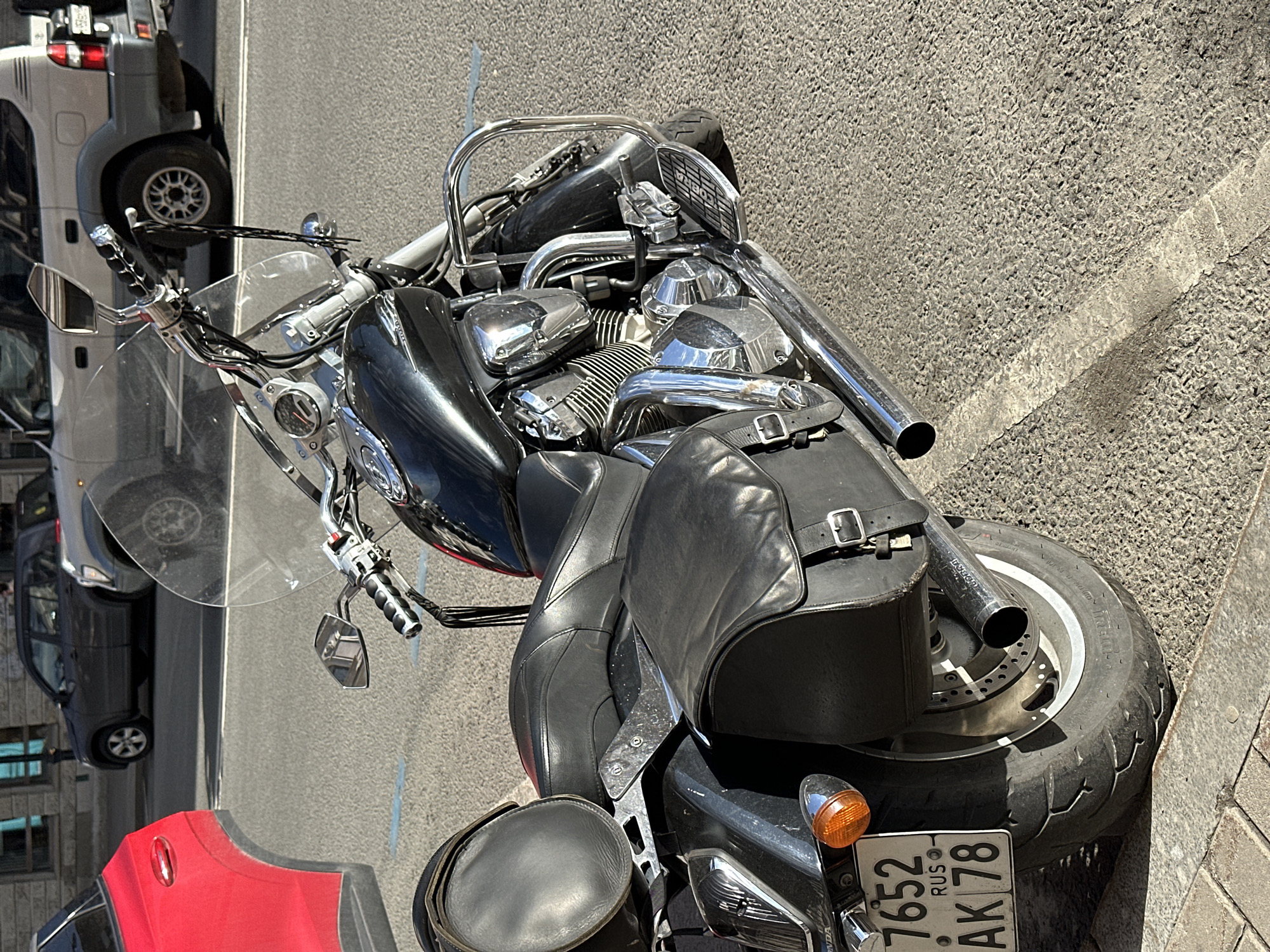 The image shows a parked motorcycle in Saint Petersburg, Russia. The motorcycle is a cruiser style, black in color, and equipped with saddlebags. The license plate is visible, reading "7652 AK 78" with the "RUS" abbreviation, indicating a Russian vehicle. The motorcycle is parked on an asphalt surface near a curb. In the background, blurred cars are visible, suggesting a street setting. The photo was taken in daylight.