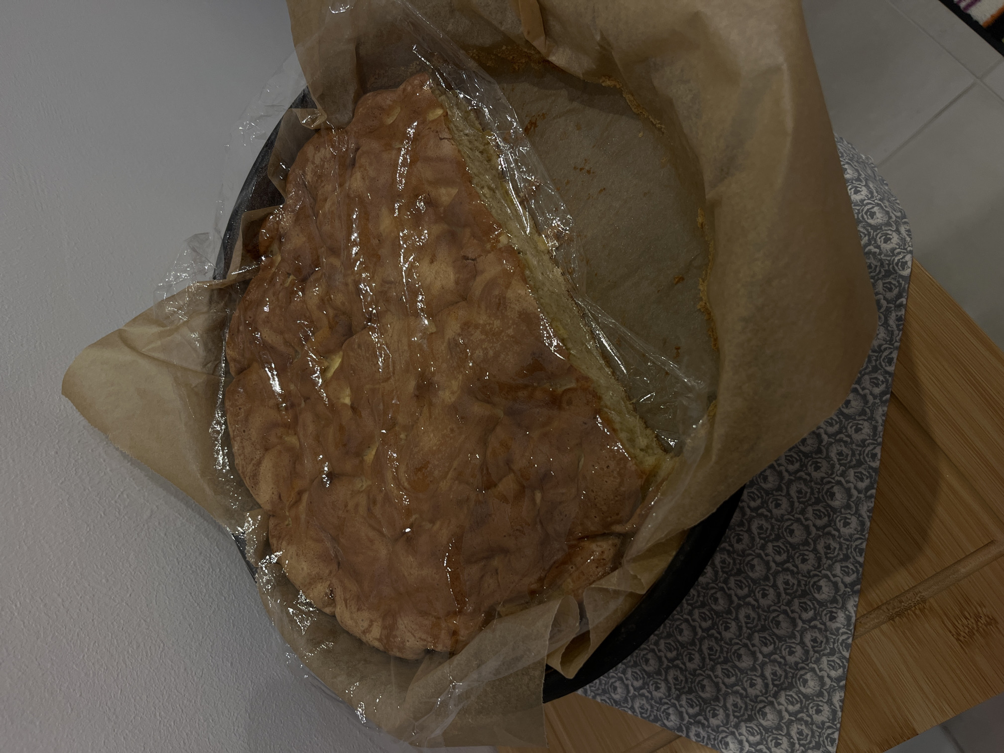 A baked apple pie in a round black baking dish is set on a wooden cutting board, with the dish lined with parchment paper and covered with plastic wrap. The pie is slightly cracked on top. A patterned gray and white cloth napkin is placed beneath the dish. The scene appears to be indoors, with a gray wall in the background on the left. There is no immediate evidence of location in Marten, Bulgaria.