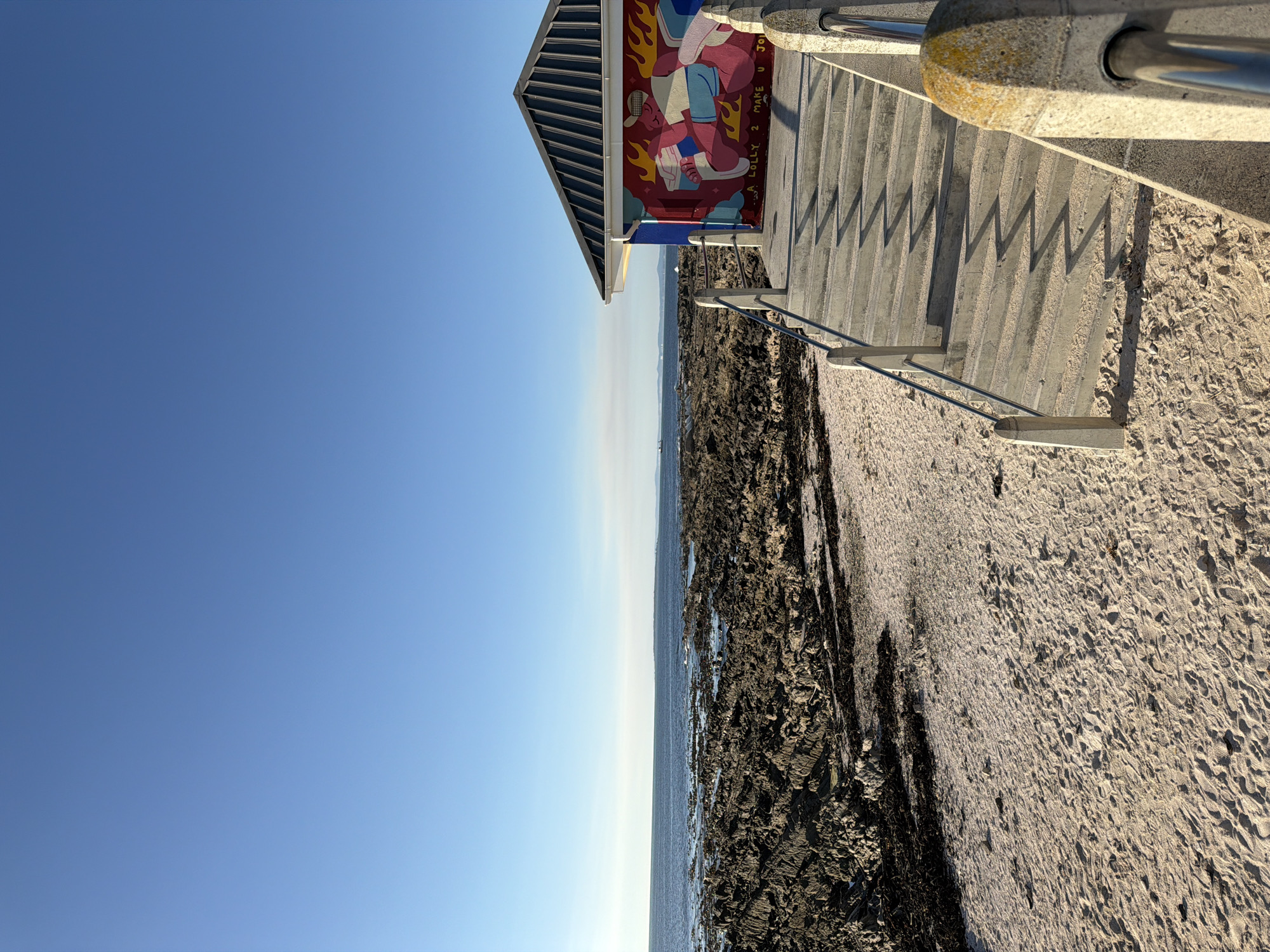 The photograph captures a view from a beach in Cape Town, South Africa. The clear blue sky dominates the left side of the frame. Along the horizon, a narrow strip of ocean meets the sky.
To the right, the image features a coastal scene with a rocky shoreline transitioning to a sandy beach. A concrete staircase with metal railings leads down to the beach. Adjacent to the staircase is a small building adorned with a colorful mural depicting figures and flames. The artwork adds a distinct visual element to the scene.