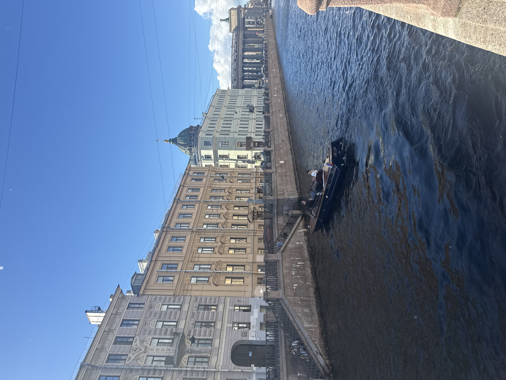 The image depicts a wide canal in Saint Petersburg, Russia, with ornate, historic buildings lining the left bank. The canal is filled with dark blue water that reflects the bright blue sky and scattered white clouds. A small, dark boat with a few passengers is moving away from the viewer on the canal. The buildings feature varied architectural styles, with multiple stories, numerous windows, and decorative facades. The buildings include a prominent structure with a green dome and a classical colonnade in the distance. A stone embankment with stairs leading down to the water is visible along the canal's edge. The overall scene captures a sunny day in the city, with the water and architecture suggesting a European urban environment.