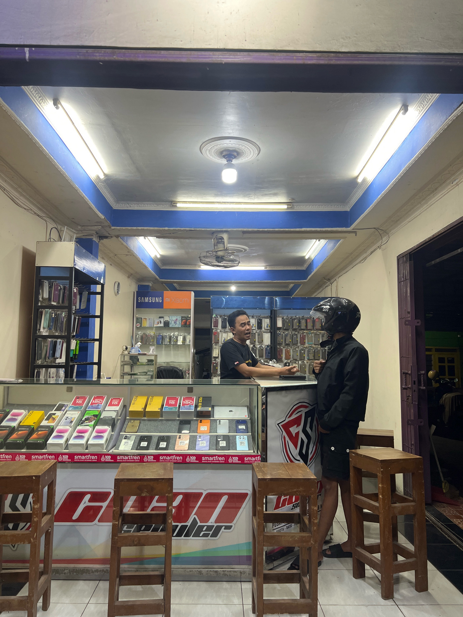 The image captures a dimly lit mobile phone shop in Kecamatan Pati, Indonesia. Two men are present. One man, wearing a black t-shirt and speaking animatedly, stands behind a counter. The other man, dressed in a black jacket and black shorts, and wearing a black helmet, is facing the counter, seemingly engaged in a transaction. The shop's interior is filled with display shelves showcasing mobile phone accessories and boxes of mobile phones. The shop has a blue and white ceiling with fluorescent lighting. Two wooden stools are placed in front of the counter. The overall impression is a small, functional retail space.