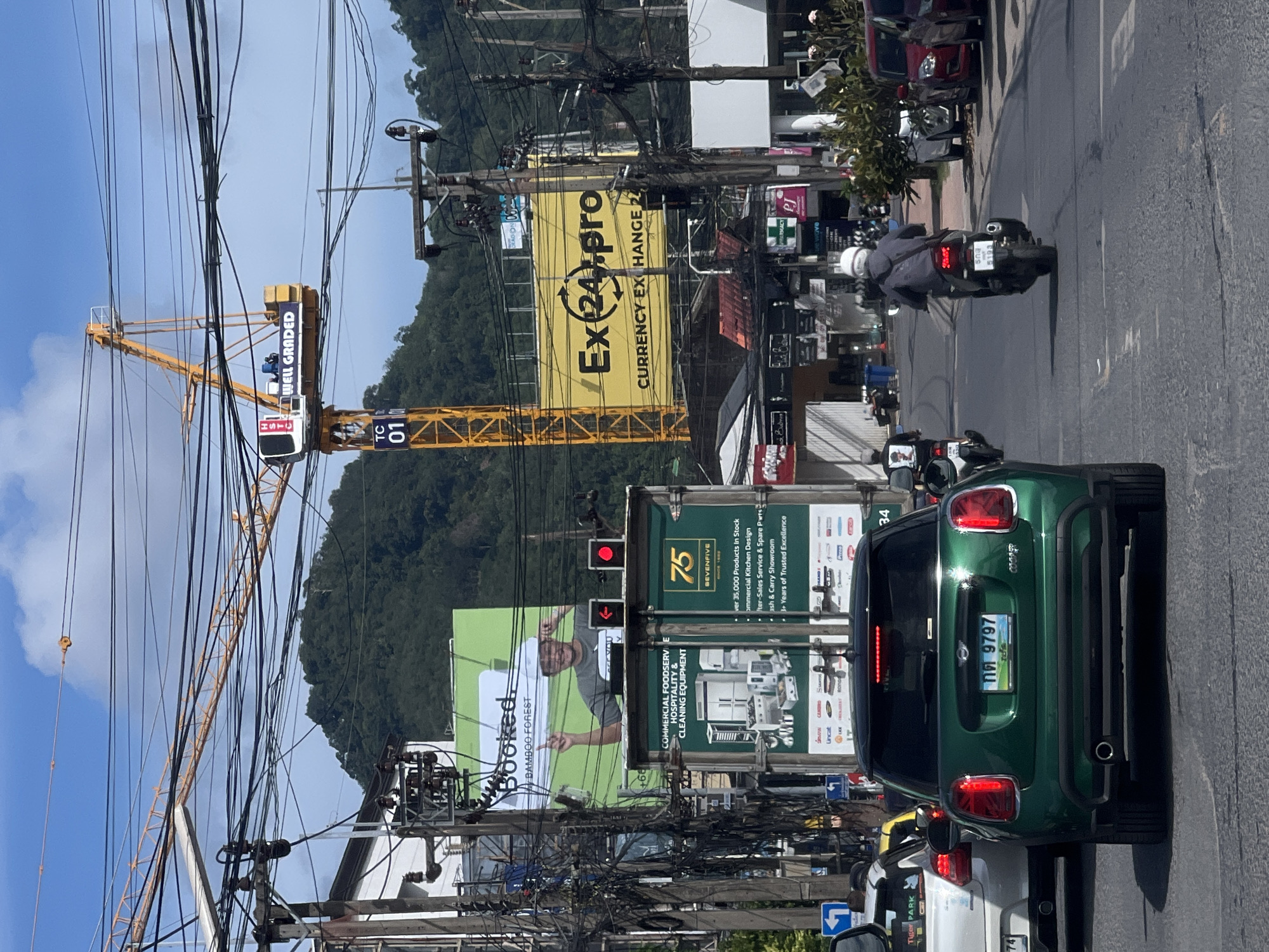 In a city scene, a green Mini Cooper is stopped in traffic. The license plate reads "กต 9797". The car is facing away from the camera. In front of the Mini Cooper, a white car is also stopped. In the background, a yellow construction crane is visible, with the words "WELL GRADED" displayed on it. A large yellow billboard with the text "Ex24.pro CURRENCY EXCHANGE 24" is also present. Traffic lights are red. The sky is blue with scattered clouds. Numerous power lines are strung across the scene. A motorcycle with a rider wearing a helmet is in the lane to the right of the Mini Cooper, also stopped. Further in the background, a lush green mountain looms.