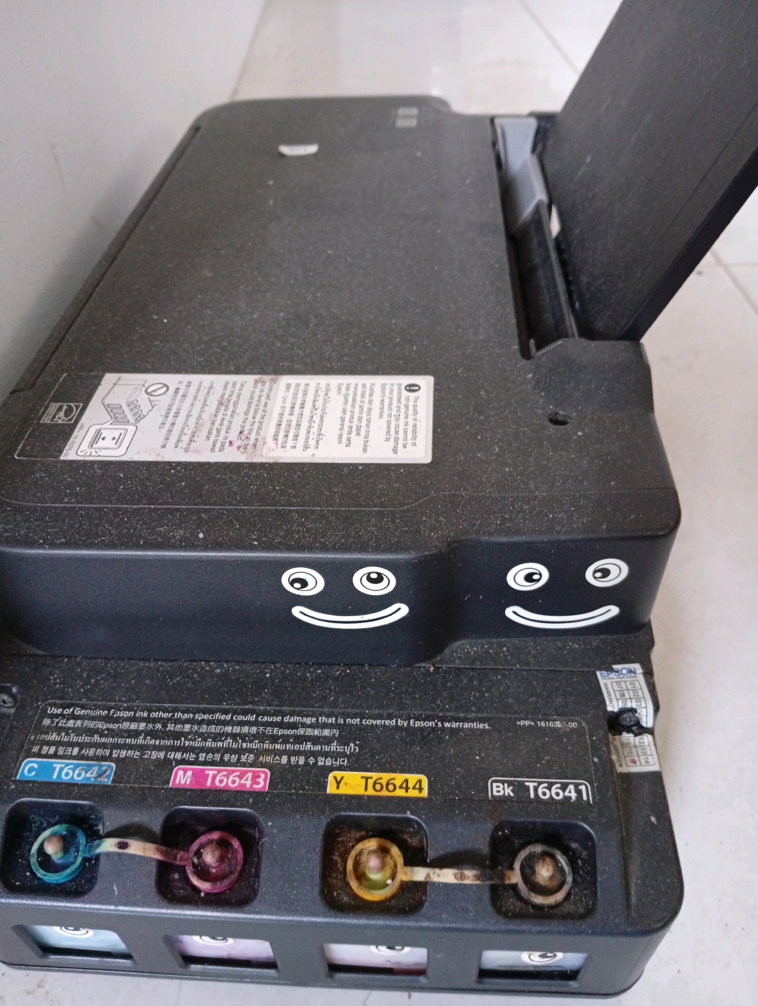 An indoor, eye-level, medium shot captures a dusty black Epson printer with two cartoon-like faces drawn on its side. The printer has its scanner lid open, revealing the interior. Below the scanner lid, there are printed labels in multiple languages, one of which states "Use of Genuine Epson ink other than specified could cause damage that is not covered by Epson's warranties." To the right of the scanner lid, a portion of a white wall is visible. At the bottom of the image, a row of ink cartridges is visible, with the labels "C T6642", "M T6643", "Y T6644", and "Bk T6641" indicating their colors and model numbers. The ink cartridges are slightly open, with remnants of colored ink visible. The printer is placed on a light-colored tiled floor. The scene is devoid of people and any specific environmental markers that would indicate a location in Kebondalem, Indonesia.