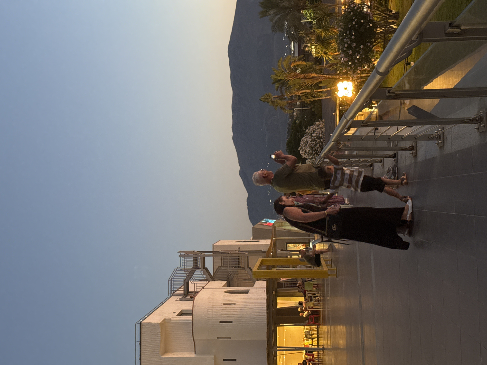An exterior, eye-level, vertical, medium shot captures a twilight scene on a paved outdoor walkway, likely in Isola delle Femmine, Italy, overlooking a town and mountains. In the foreground, a woman in a long black dress and white shoes stands with her back to the camera, facing left. To her right, a young boy in a striped shirt and dark shorts walks away from the camera, his right arm raised. To the right of the boy, a man in a green shirt and glasses is captured in profile, facing left and holding a cell phone up to take a picture. He appears to be standing on a metal railing that borders the walkway. Behind the railing is a grassy area with palm trees and bushes, illuminated by warm, ambient lights, including a large, glowing orb light fixture. Further back, a multi-story white building with a curved facade and an external metal staircase is visible, with illuminated windows and outdoor seating areas. In the distance, dark mountains form the horizon against a pale blue sky.