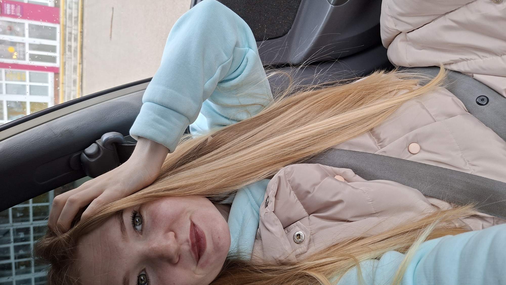 A young woman with long blonde hair is sitting in what appears to be a car, secured by a seatbelt. She is wearing a light blue sweatshirt and a light pink puffer jacket. She has green eyes and fair skin. Her right hand is raised, with fingers touching her head. Outside the window, a building with a pink facade and multiple windows is visible. The presence of water droplets on the window suggests it might be raining. Given the location of Chaikovsky, Russia, is specified, it suggests this scene is taking place within or near that city.