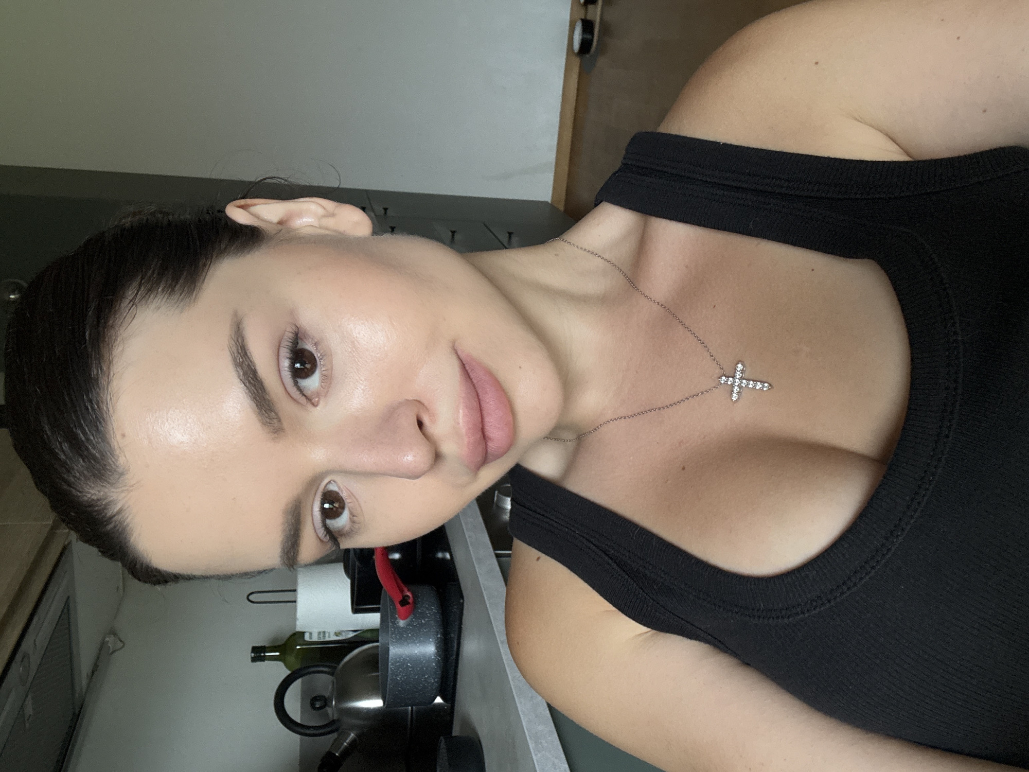 A woman with dark hair pulled back is photographed from the chest up. She is wearing a black tank top and a silver necklace with a cross pendant. Her makeup is natural with a focus on radiant skin and neutral lip color. The background appears to be a kitchen area, with a stove, pots, and a kettle visible. The overall lighting suggests an indoor setting during the daytime. The image was taken at a residence in Prague, Czechia.
