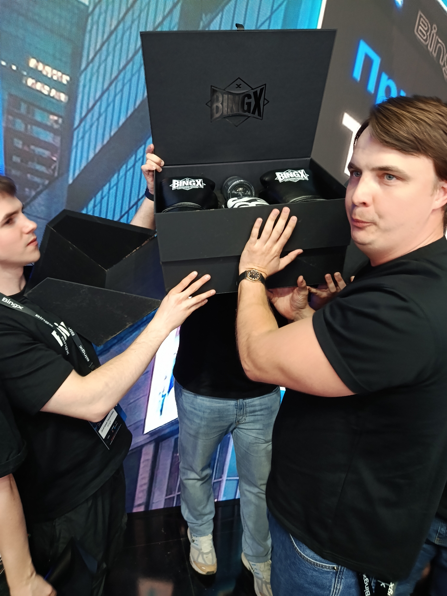 The image depicts three people holding a black BingX-branded box. The person on the right is a fair-skinned man with brown hair, wearing a black t-shirt, blue jeans, and a wristwatch. The person on the left is a fair-skinned young man with dark hair, wearing a black t-shirt with a BingX lanyard around his neck, and holding the open side of the box. The third person, mostly obscured in the center, is wearing a black shirt, light blue jeans, and white sneakers. The box contains items labeled with the BingX logo. A large screen in the background shows an urban cityscape and the BingX logo. The location seems to be at an event or conference, possibly in a city.