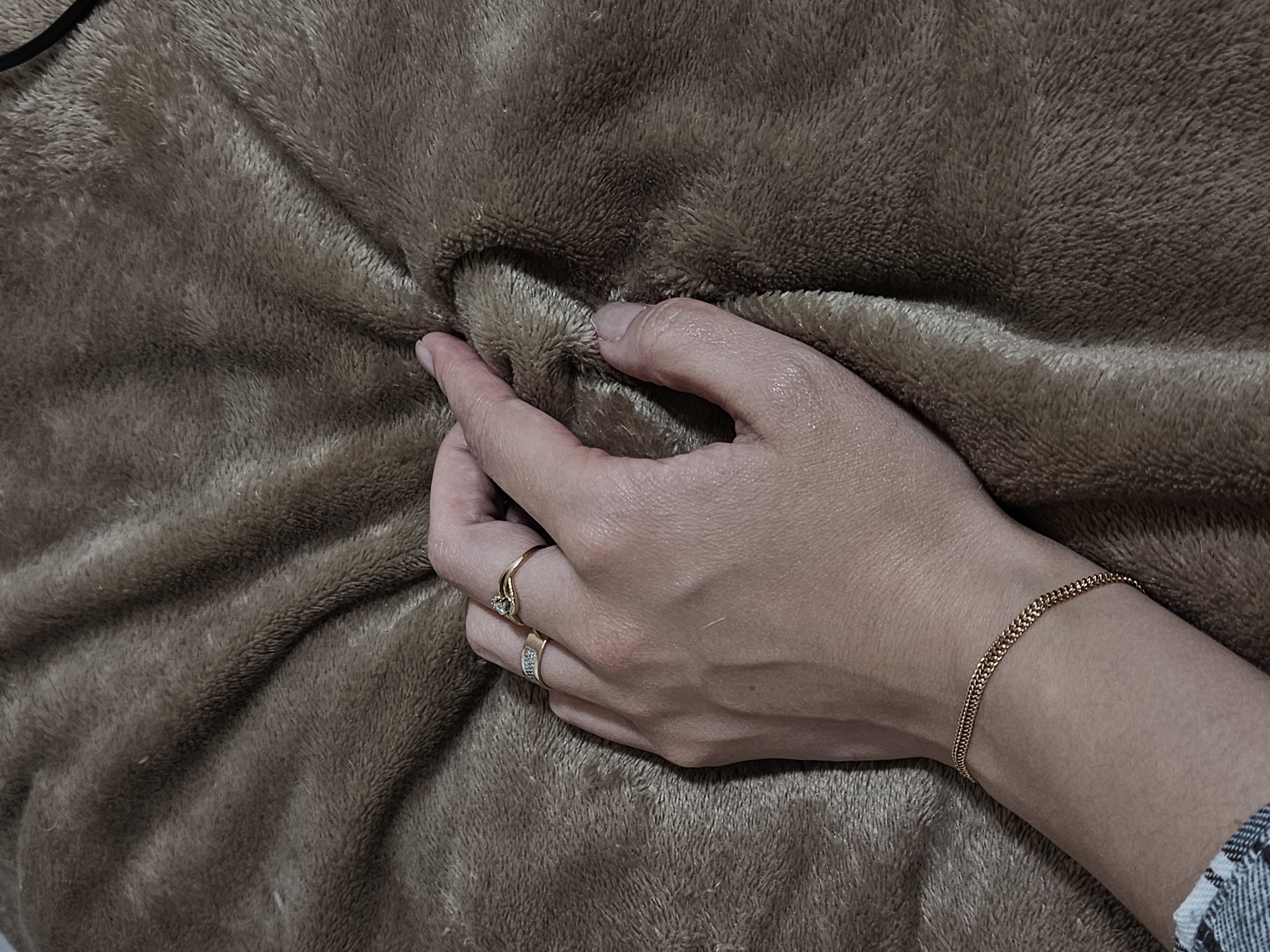 A close-up shot features a human hand gently gripping a textured, plush, brown fabric, likely a blanket or throw. The hand, appearing to be an adult's, is positioned in the center-right of the frame, with fingers creating soft indentations in the material.
The skin of the hand appears fair, with visible veins on the back of the hand. Two rings adorn the fingers: a gold-colored ring with a small, light-colored stone on the ring finger, and a gold-colored band with tiny clear stones on the middle finger. A delicate, rose gold-toned chain bracelet with a braided or double-strand design is worn on the wrist. A small portion of a patterned sleeve, possibly plaid or checkered in shades of blue, white, and grey, is visible near the wrist on the far right.
The fabric itself is the dominant background element, filling the entire frame. Its texture is soft and fuzzy, characterized by short, dense fibers that create varying shades of brown and light reflections, particularly where the hand compresses it. The folds and creases in the material suggest its pliable nature. The lighting is soft, highlighting the tactile qualities of both the skin and the fabric. There is no visible text in the image. The scene conveys a sense of comfort and a focus on sensory experience.