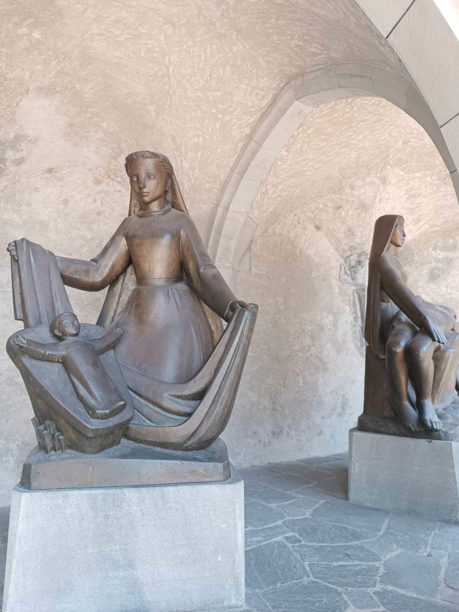 This image depicts two bronze sculptures of the Virgin Mary and the infant Jesus, located in Vaduz, Liechtenstein. The sculpture in the foreground shows the Virgin Mary holding a veil that drapes over a cradle containing the infant Jesus. The infant is positioned in a supine pose with arms outstretched. The sculpture in the background portrays the Virgin Mary seated and holding the infant Jesus in her lap. Both figures are rendered in a stylized, modern artistic fashion, with smooth surfaces and simplified features. The sculptures are placed on stone pedestals against a backdrop of an arched stone structure with a textured, light-colored wall and a cobblestone floor. The lighting suggests an outdoor, possibly shaded, environment.
