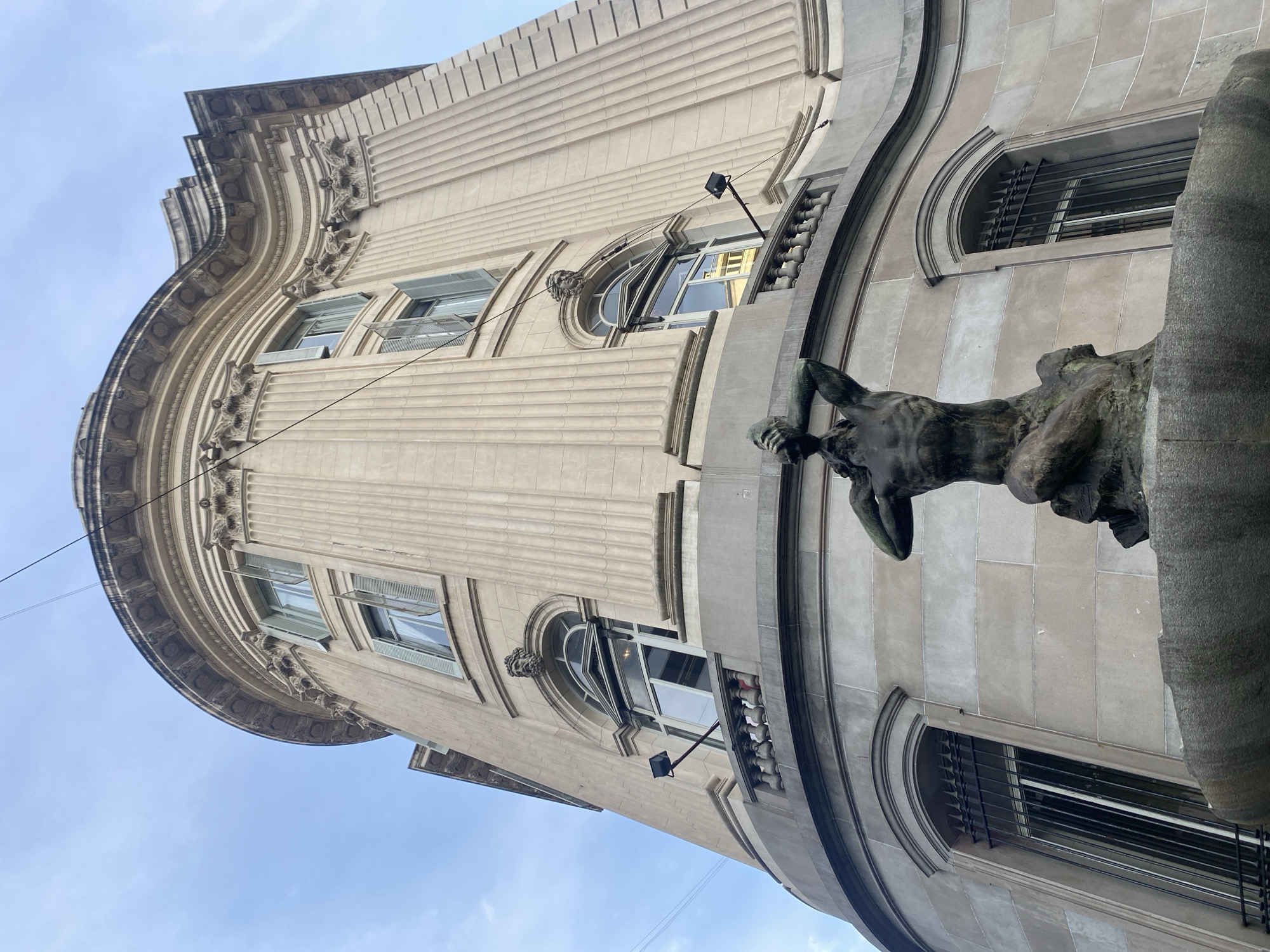 An upward-angled, medium shot captures a portion of a building's exterior. The building features classical architectural elements, including fluted columns, ornate detailing, and arched windows. A bronze statue of a human figure clings to the facade on the right side of the frame. The building is in Buenos Aires, Argentina.