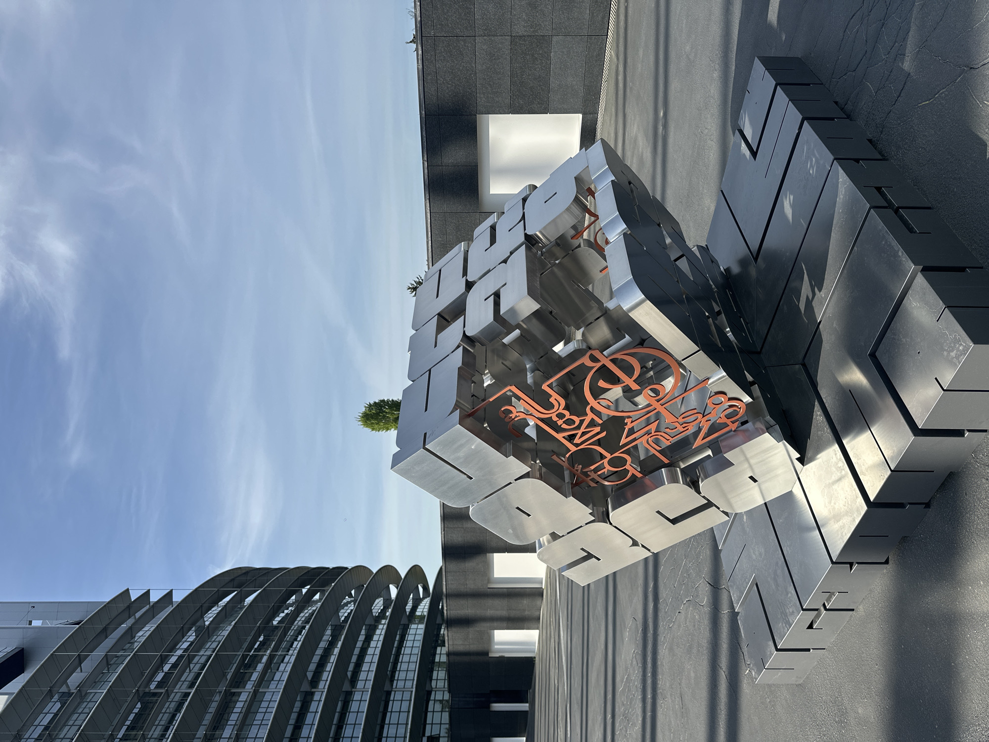 The image captures a modern urban landscape in Strasbourg, France, under a clear blue sky dotted with wispy white clouds. The central subject is a large, metallic sculptural installation shaped like a deconstructed cube, composed of three-dimensional letters. The primary visible letters on the sculpture are silver or chrome-finished, forming parts of words, with "ULTURES" clearly discernible on one face, likely part of "CULTURES" or "FUTURES." An intricate, stylized script or logo in a contrasting copper or bronze color is also visible on another face of the sculpture. Strong sunlight casts sharp, dark shadows from the sculpture onto the dark grey ground, suggesting a clear mid-day setting.
In the background, two distinct modern buildings frame the sculpture. To the left, a building features an architectural design with multiple large, curved, arched sections of glass and metal, forming a unique, wave-like facade. To the right, another structure presents a dark grey, possibly tiled or paneled, exterior with a prominent bright white rectangular opening or illuminated section. A small, distant tree is visible on a rooftop. No people or specific activities are depicted, rendering the scene static and emphasizing the architectural and artistic elements.