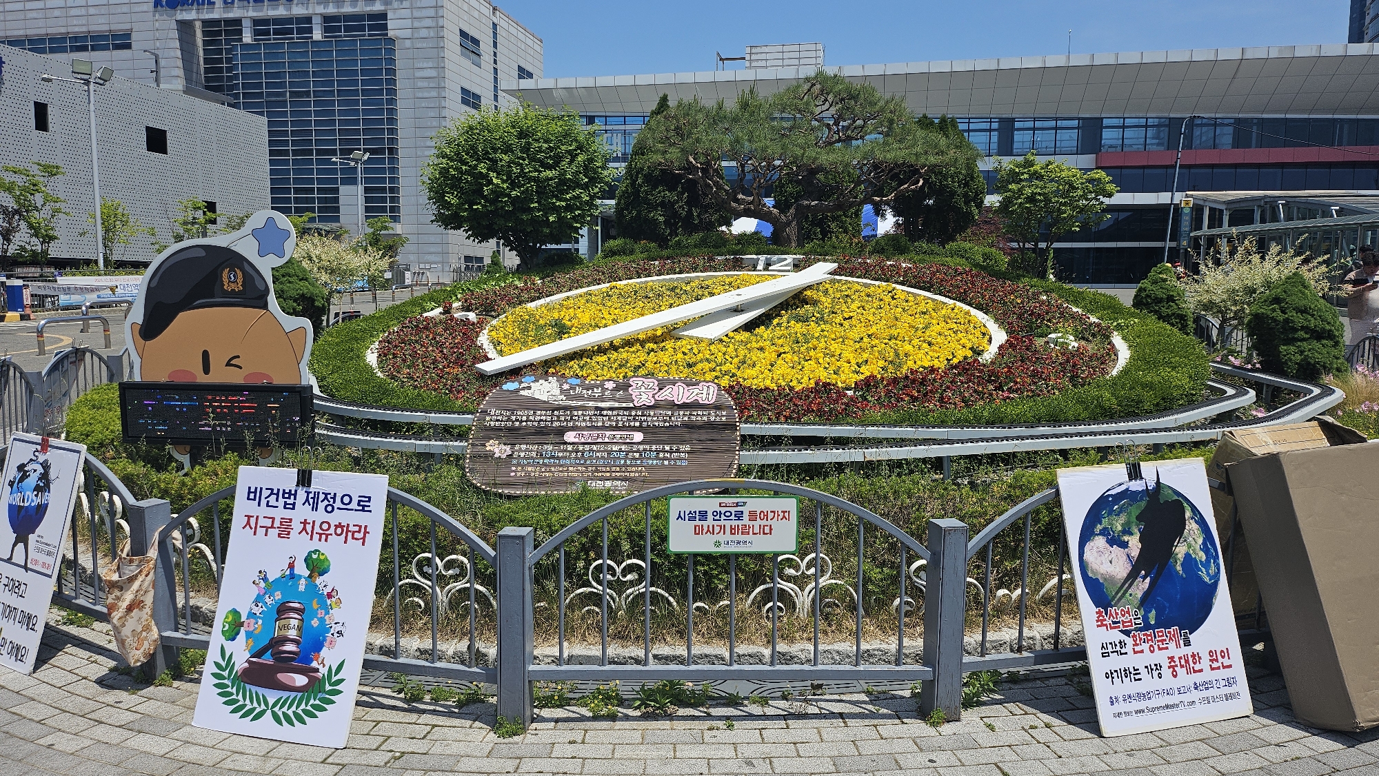 Here is a detailed description of the image:
The image shows a flower clock display in Daejeon, South Korea. The clock face is made of yellow and red flowers, with white clock hands. There is a sign in the center of the clock face written in Korean. Behind the clock is a manicured green landscape. In the background, there are modern buildings with a clear blue sky overhead.
In front of the flower clock is a decorative metal fence. Attached to the fence are several signs with Korean text. These signs seem to be part of a protest or public message display. A cartoon character cutout is displayed on the left side of the flower clock. There is a small train track surrounding the flower display. The ground is paved with brick.