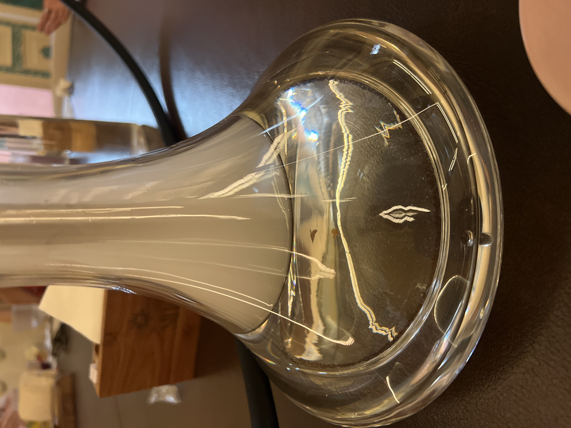 A close-up shot of a glass vase with a bulbous base and a narrow neck. The vase is resting on a dark, possibly brown, textured surface. The inside of the vase appears to be filled with a white, opaque substance, possibly liquid or vapor. Reflected light creates bright streaks and highlights on the vase's surface. A small pink object is partially visible in the upper right corner. Other items are reflected in the vase’s glass and visible in the background, including furniture and a wooden block. The scene appears to be indoors. Location is unknown.