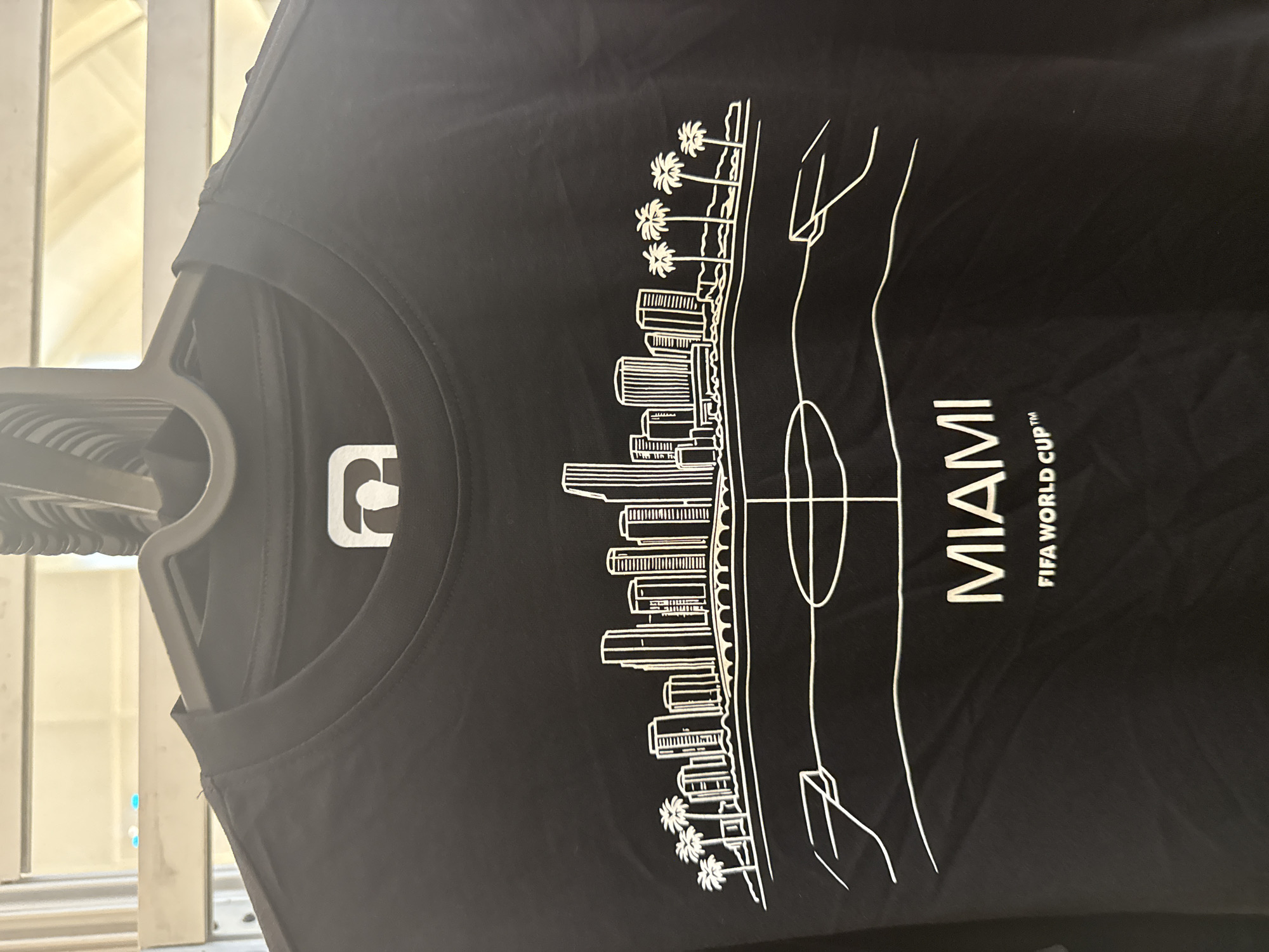 A black, long-sleeved t-shirt is displayed on a dark grey plastic clothes hanger, viewed from a slightly elevated frontal perspective within an indoor setting in Sunrise, United States of America. The t-shirt's ribbed crew neck shows a white brand tag with a stylized dark grey logo, resembling an abstract 'P' or '6'. The garment features a prominent white outline graphic depicting a cityscape, specifically Miami, with multiple tall buildings, a bridge, and groups of palm trees flanking the skyline. Below the cityscape, a stylized football (soccer) pitch outline is present, showing the center circle, halfway line, and partial goal areas. Beneath this graphic, two lines of white text are centered: "MIAMI" in large uppercase letters, followed by "FIFA WORLD CUP™" in smaller uppercase text. The t-shirt fabric shows minor creasing. The background is out of focus, revealing horizontal light-colored elements consistent with blinds or shelving, with indistinct light sources and two small blue illuminated points in the upper left quadrant.