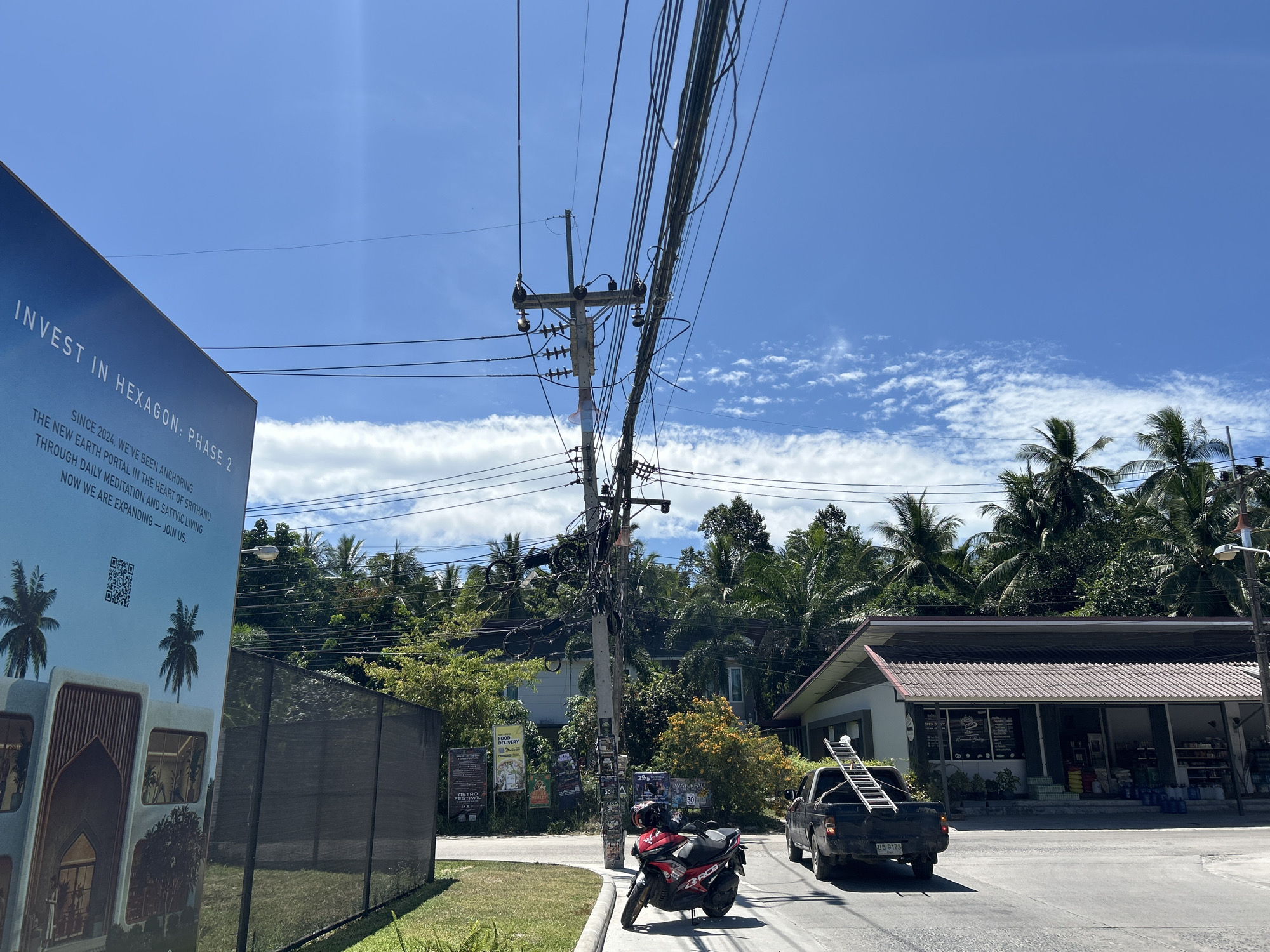The image depicts a street scene in ตำบลเกาะพะงัน, Thailand on a bright, sunny day. A sign advertising real estate ("INVEST IN HEXAGON: PHASE 2") stands in the left foreground. A red and black motorcycle is parked near a utility pole laden with wires. A black pickup truck, carrying a ladder, drives on the asphalt road in front of a single-story building with a tiled roof and shop-front windows. Tropical vegetation, including palm trees, lines the background. Various posters are affixed to the utility pole and nearby wall. The sky is blue with scattered clouds.