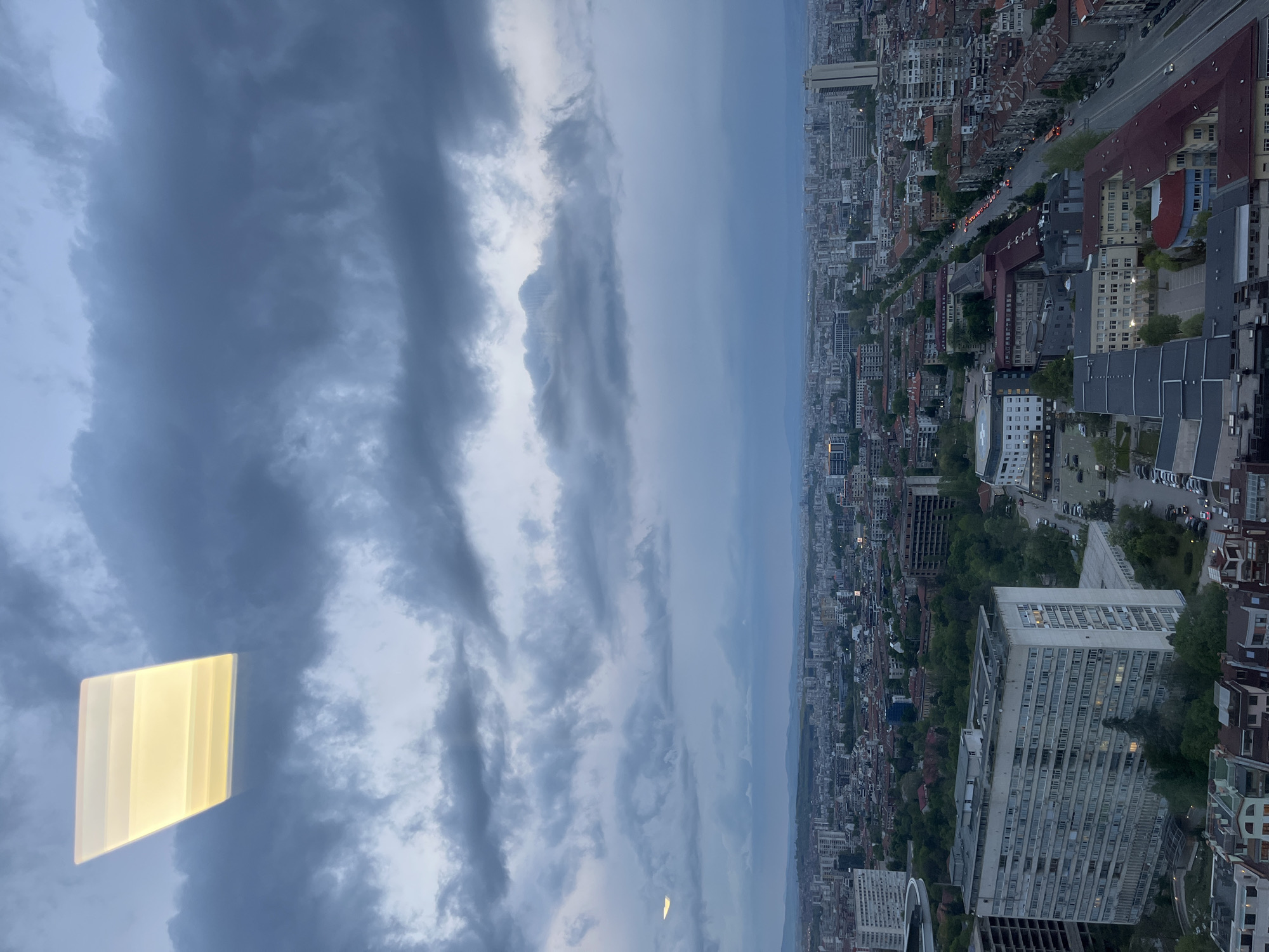 This high-angle, wide-view photograph captures a vast cityscape, identified as Sofia, Bulgaria, under a dramatic, heavily clouded sky. The image appears to be taken from a tall building or observation point, looking down and across the urban expanse.
The upper portion of the scene is dominated by dense, dark grey, elongated clouds, suggesting overcast weather, possibly during dusk or a very gloomy day. Patches of lighter sky are visible between the imposing cloud formations.
Below the horizon, the city stretches out extensively, with countless buildings of varying heights and architectural styles. A mix of older structures with red-tiled roofs and modern, multi-story buildings, some appearing residential and others commercial, populate the landscape. Green areas and tree clusters are interspersed throughout the urban fabric. Roads are visible winding through the city, with faint streaks of vehicle lights indicating ongoing traffic. Notable specific buildings include a tall, light-colored structure with many windows in the mid-ground, and another large, white, rectangular building in the foreground on the right.
A prominent detail in the upper-left of the image is a bright, rectangular, yellow-orange reflection, likely an interior light source reflecting off the window through which the photo was taken. A smaller, similar reflection appears further to the right. The overall lighting is dim, consistent with either a late-day scene or heavy cloud cover. No discernible text is visible within the image.