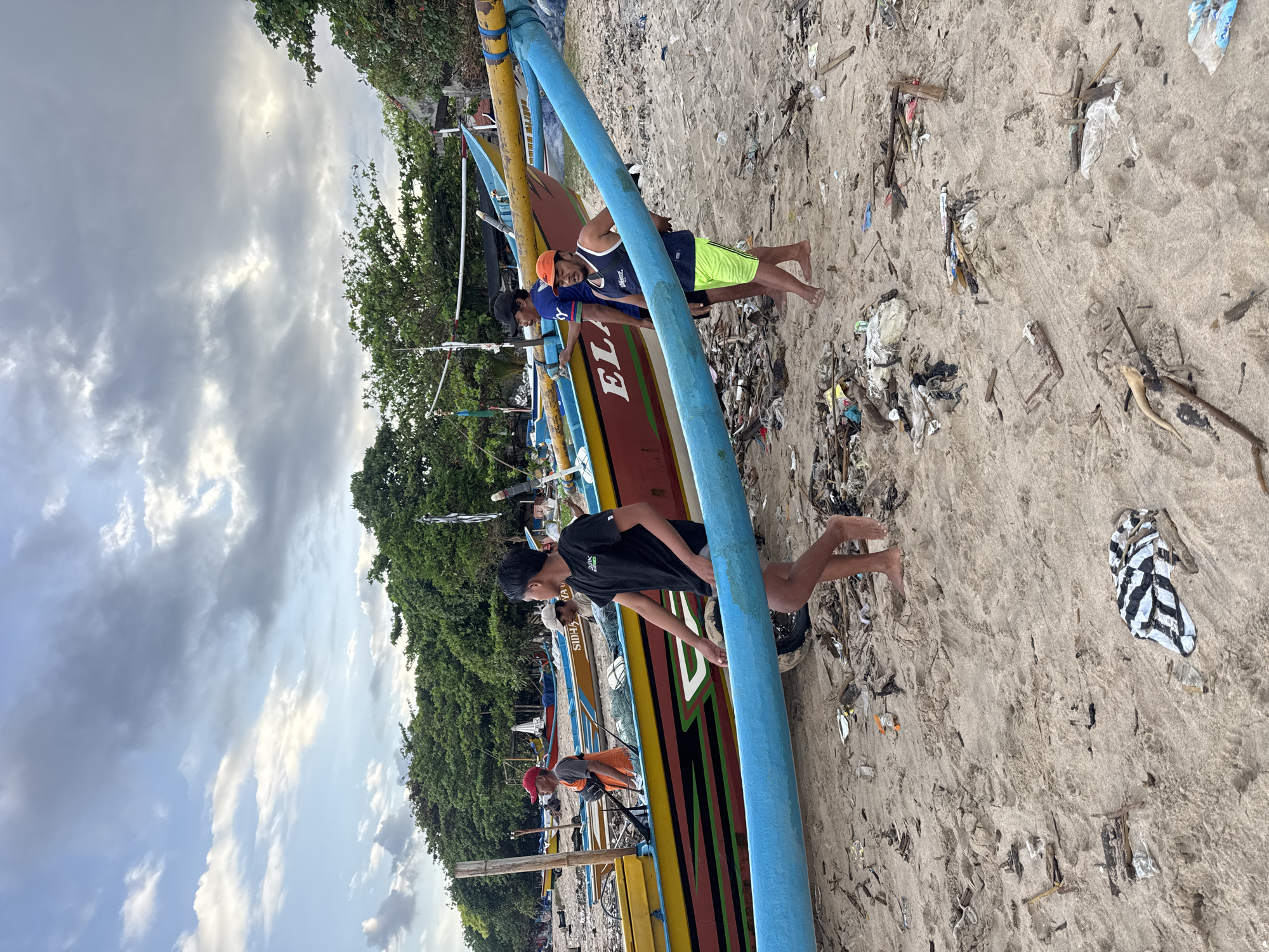 The image captures a scene on a beach in Kuta Selatan, Indonesia. Several traditional outrigger boats, painted in vibrant colors, are beached on the sand. Litter and debris are scattered across the sandy surface. Three individuals are visible near the boats: one wearing a black shirt, another in a blue tank top and yellow shorts, and a third partially visible wearing blue. They appear to be interacting with the boats. The sky is overcast with a mix of clouds. Lush green vegetation forms the backdrop to the beach scene.