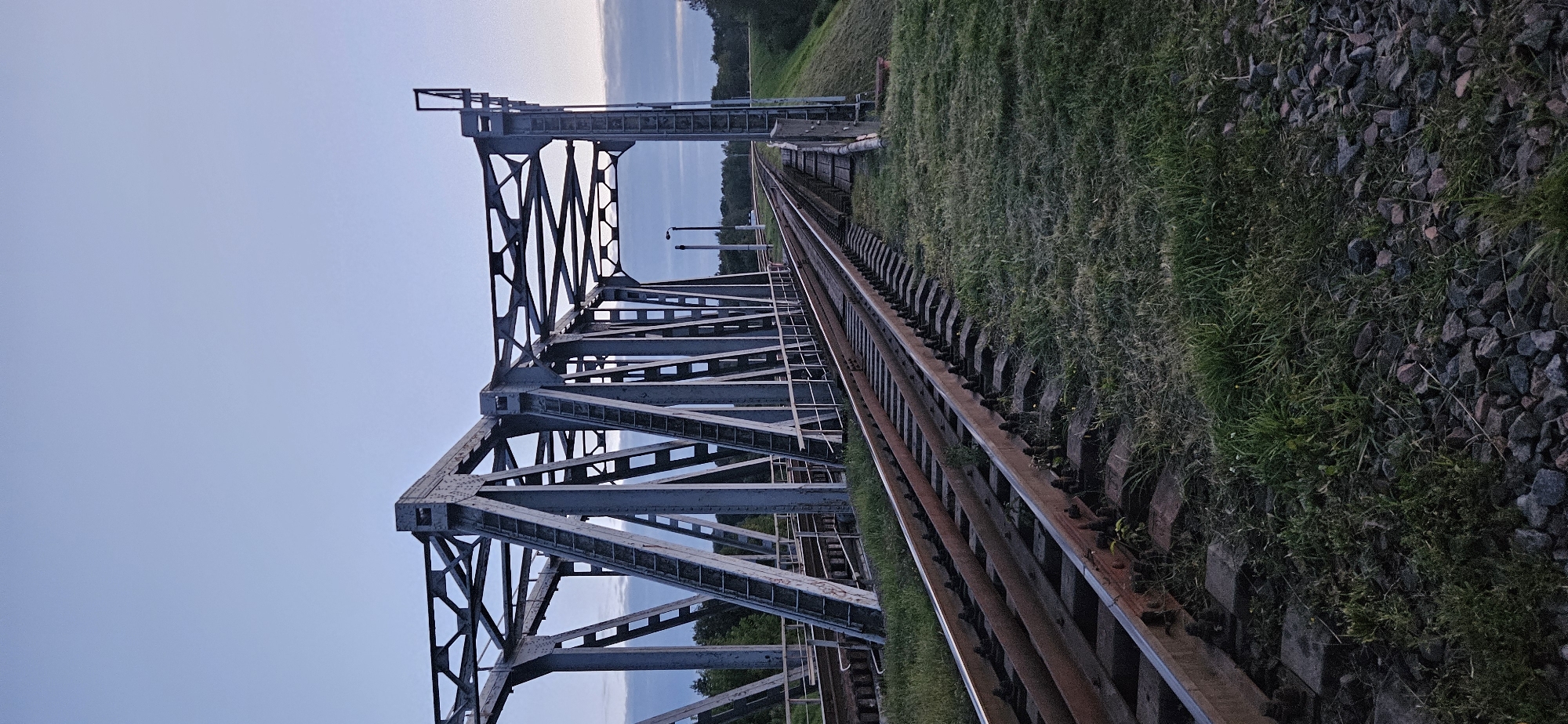 This is a vertical, medium-shot, eye-level photograph of a railway bridge at dusk in Vitsebsk, Belarus. The bridge is constructed of grey metal trusses and carries two sets of railway tracks. The tracks are rusty and appear to be made of metal rails supported by concrete sleepers. The tracks curve away from the viewer and disappear into the distance, leading across the bridge and into a wooded area. The sky is a pale blue with some scattered clouds. The ground on either side of the tracks is covered in green grass and dark grey rocks. There are no people or vehicles visible in the image. The scene is quiet and still, suggesting a rural or suburban setting.
