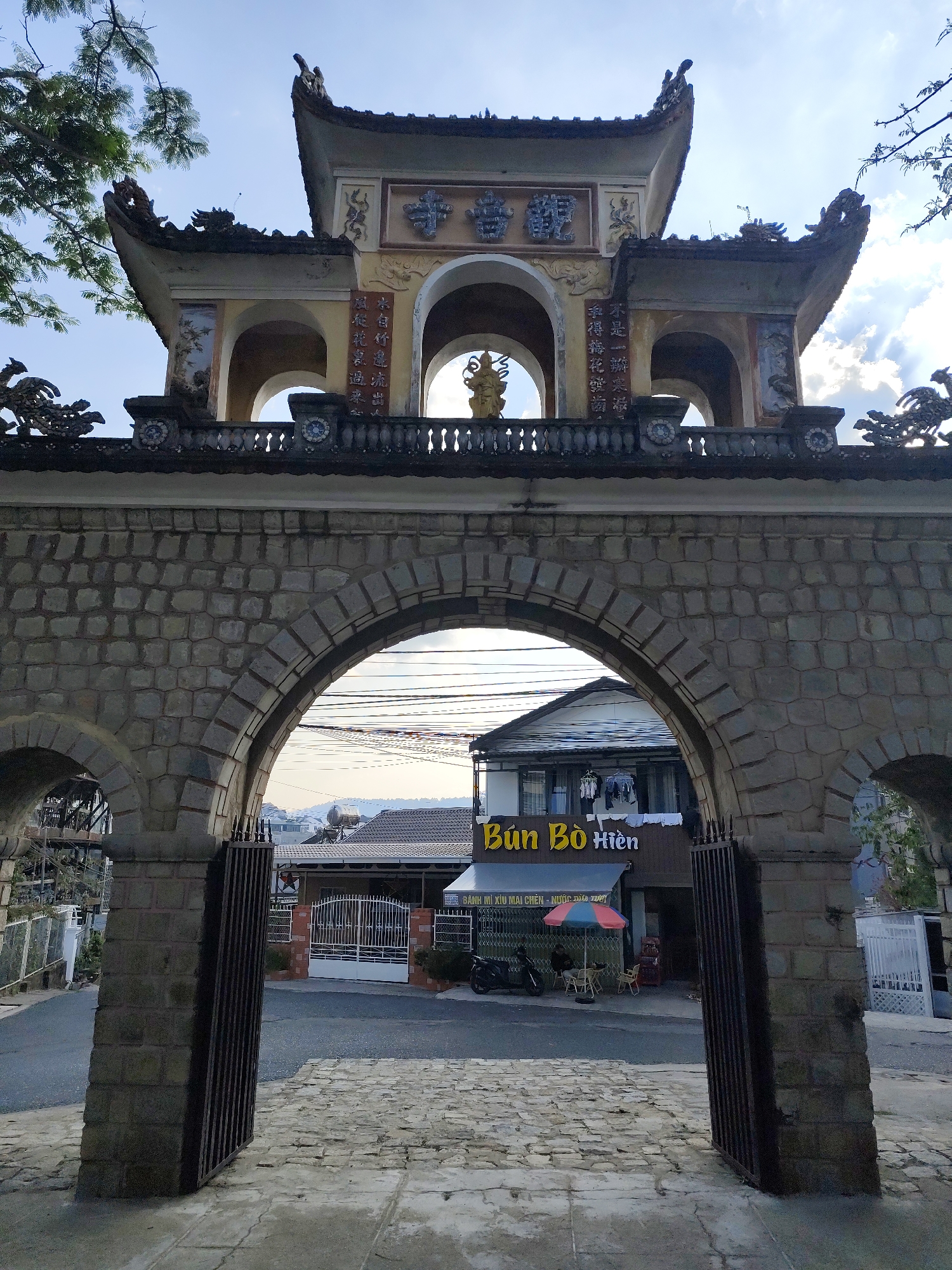 The image is a view through the stone archway of the "Tran Quoc Toan" gate in Da Lat, Vietnam, looking out onto a street. The archway is made of roughly hewn stones. Above the main arch is a smaller arched structure, ornately decorated with Chinese calligraphy. On top of that structure sits a golden Buddha statue in the center with archways on both sides of it. Beyond the gate, across the street, is a small restaurant called "Bún Bò Hiền" with its name displayed in yellow lettering. A red-and-blue striped umbrella stands outside the restaurant, with a couple of chairs and tables beneath it. A motorcycle is parked nearby. In the background, rooftops and hills can be seen under a slightly overcast sky. The gate's metal doors are partially open.