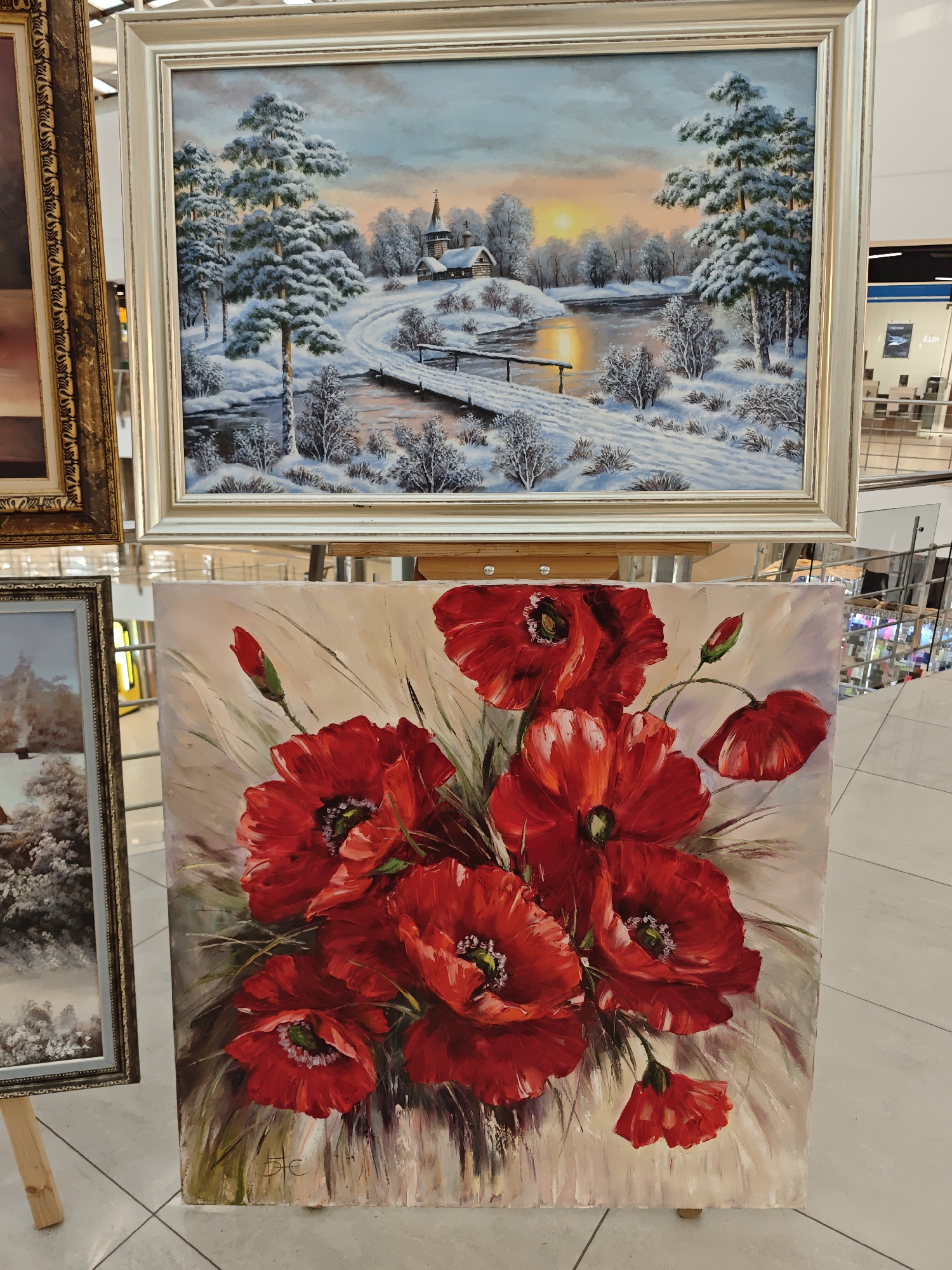 The image shows two paintings displayed in what appears to be a shopping mall in Dmitrov, Russia. The foreground painting is a close-up of bright red poppies, with a few unopened buds, painted on a beige background. The artist's signature is visible in the bottom left corner. Behind this, there's a framed winter landscape painting depicting a snow-covered scene with a church and bridge over a river at sunset. Other framed paintings are partially visible on either side. The scene is set on a tiled floor in an area with visible escalators and shops in the background, suggesting the paintings are on display for sale or exhibition in the mall. There are no people visible in the immediate foreground.
