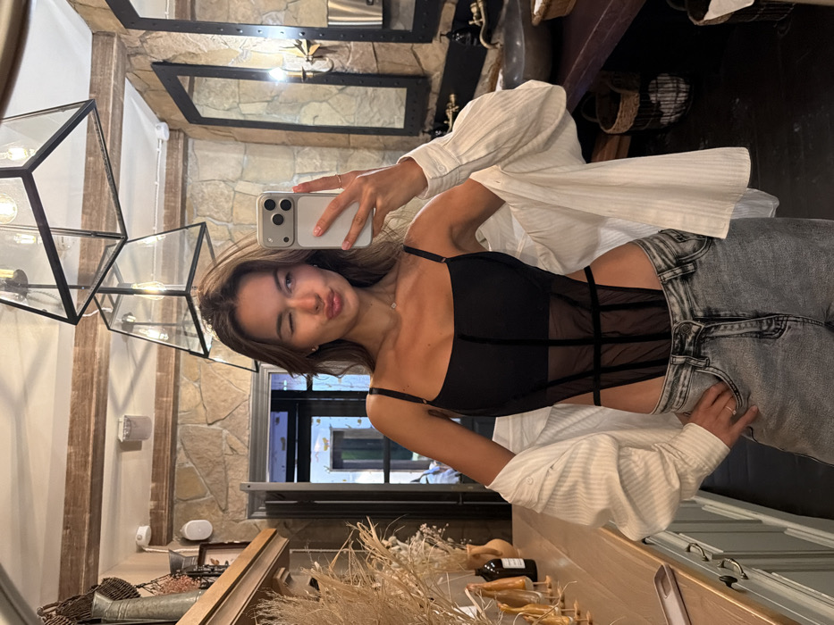 An interior scene within a public establishment in Krasnoyarsk, Russia, features a young adult female capturing a mirror selfie. The woman, with long brown hair, is positioned centrally, winking her right eye and puckering her lips. She is wearing a black, strappy, corset-style bodysuit with sheer mesh panels, over which a white, unbuttoned, long-sleeved shirt is loosely draped. Light-wash denim jeans complete her attire. A silver ring is visible on her right ring finger. She holds a light-colored smartphone with a prominent camera array in her left hand to take the photo, while her right hand rests on her hip.
The background reveals an environment with a rustic-modern aesthetic. The primary wall surface is composed of irregular, warm-toned stone or faux stone. To the left, a wooden shelf displays dried ornamental grasses in a metallic vessel and various small decorative objects. A dark wooden doorway is visible further left, leading to a brightly lit area. Above the shelf, a white wall-mounted speaker is present.
To the right, two large, dark-framed mirrors reflect parts of the room, including three black-framed, geometric pendant lights with exposed bulbs hanging from the ceiling. Below these mirrors, a dark, rustic-style vanity counter features a dark, possibly stone or concrete, basin sink. Dark, wall-mounted faucets and soap dispensers are visible above the sink, along with wall sconces providing additional illumination. Two woven baskets are placed on the floor beneath the vanity. The overall lighting is bright, combining artificial light from the fixtures with ambient light from the doorway.