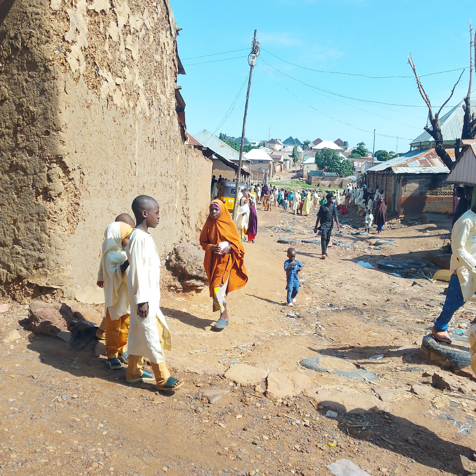This image depicts a dusty street scene in a village. The foreground is dominated by a rough, textured mud wall on the left. Several children are visible, some in traditional attire. In the center, a woman in an orange headscarf and patterned skirt walks towards the viewer, holding a white object. To her right, a young child in a blue outfit walks away. Further down the street, a procession of people, many in light-colored garments, are walking, some towards the viewer and some away. A yellow auto-rickshaw is parked on the side of the road behind some of the people. The street is unpaved and appears to have some debris and puddles of water. In the background, buildings with corrugated metal roofs and some trees are visible under a clear blue sky. A utility pole with wires and some antenna equipment stands along the street. The overall environment suggests a rural or semi-urban setting, likely in a warm climate.