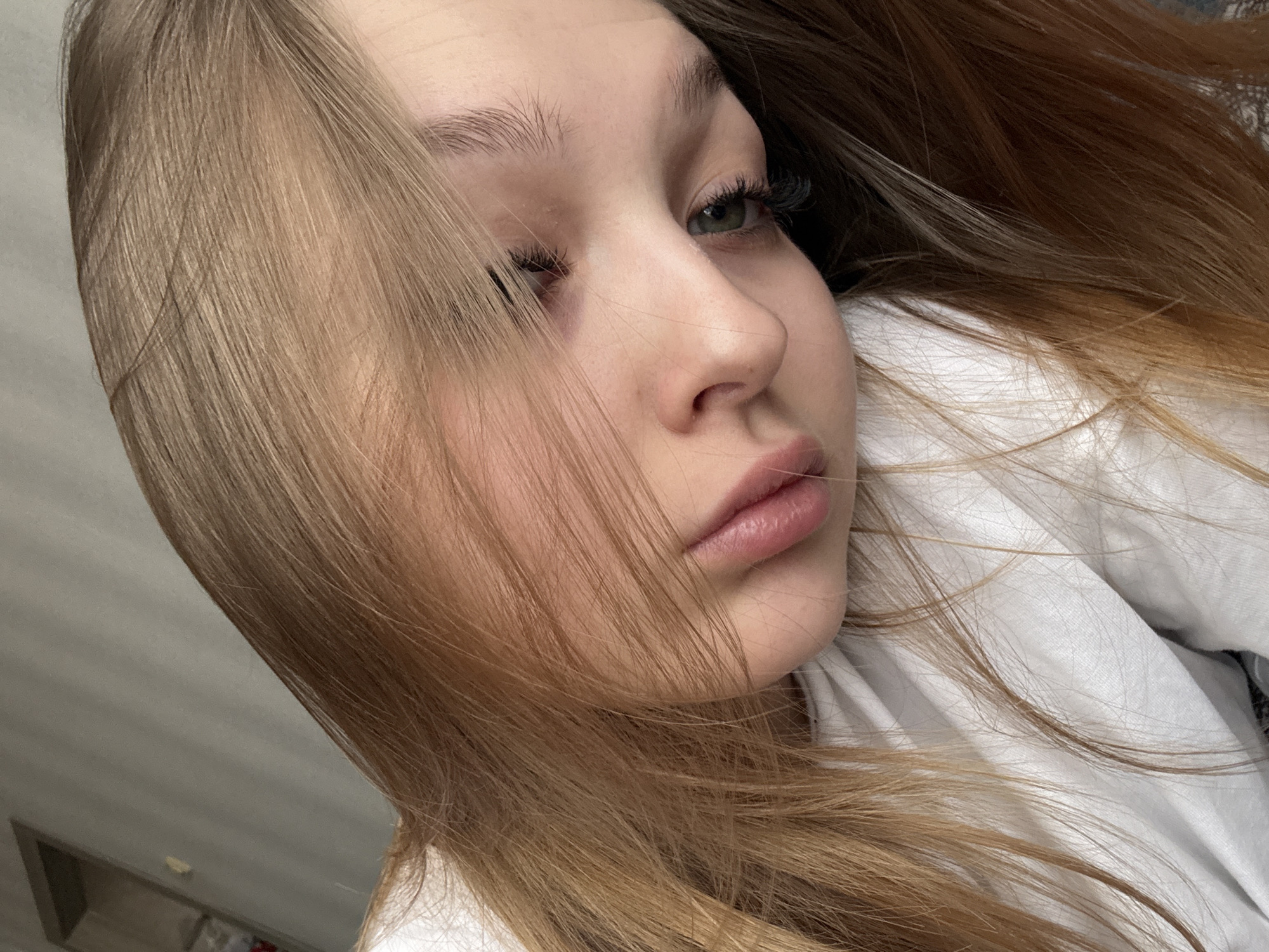 A close-up, high-angle portrait features a young person, likely female, with long, straight, light brown and blonde hair cascading around her face. Her head is tilted, and she appears to be lying down or reclining against a white fabric, possibly a bedsheet or pillow. Her light green eye, framed by prominent dark eyelashes, looks upwards and to the right, away from the camera. Her other eye is largely obscured by strands of hair. Her skin is fair, and her lips are slightly parted, showing a natural pink hue. Her expression is neutral and somewhat pensive or relaxed. A portion of her dark brown hair is visible on the right side of the frame, blending with the lighter strands. The background on the left is an out-of-focus, light gray wall or surface, suggesting an indoor setting. There are no specific location cues visible within the image, though the context provided indicates Chaikovsky, Russia. No visible text is present.